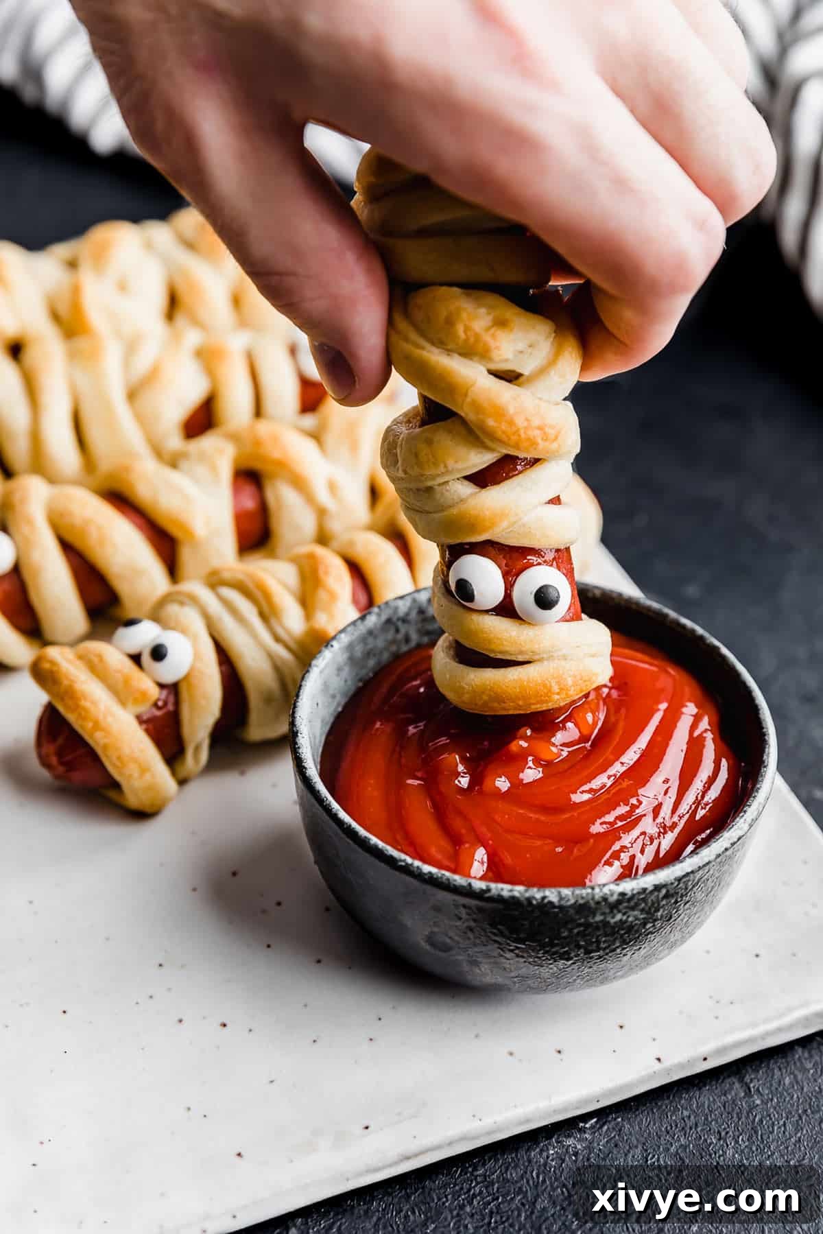 Creepy Canine Wraps 10 A hand dipping a Mummy Hot Dog into a bowl of ketchup.