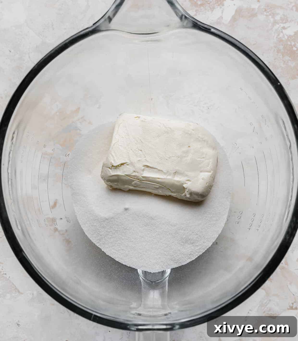 A glass mixing bowl with granulated sugar and a block of white cream cheese in it.
