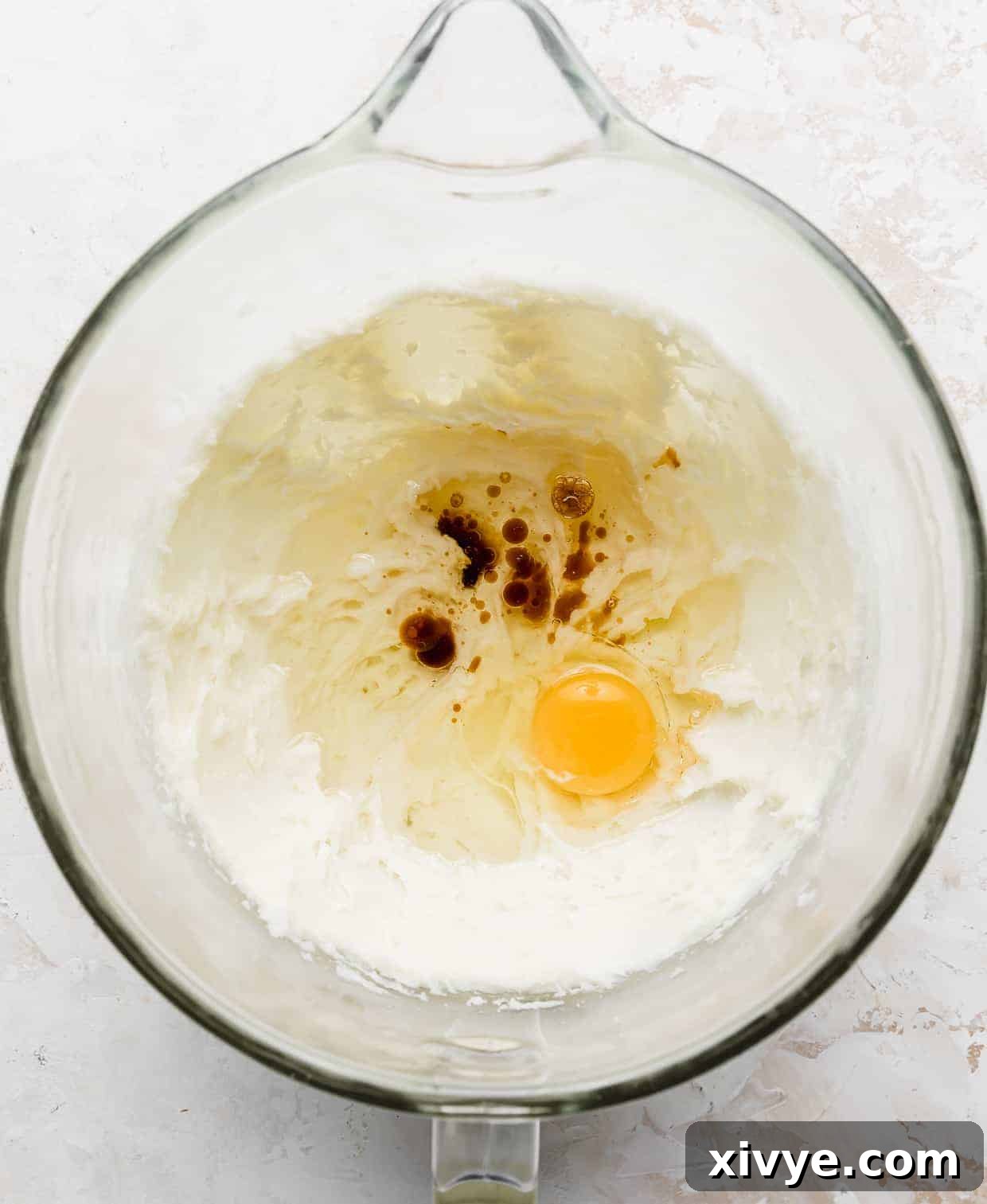 An egg, oil, and vanilla extract in the bowl of a stand mixer with creamed sugar and cream cheese.