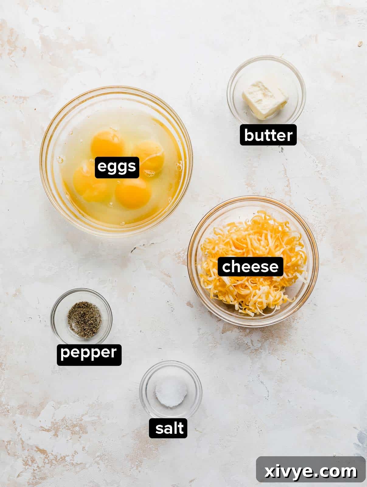 Gooey Cheese Scramble 3 Ingredients used to make Cheesy Scrambled Eggs on a textured white background, including eggs, butter, salt, pepper, and a block of cheese.