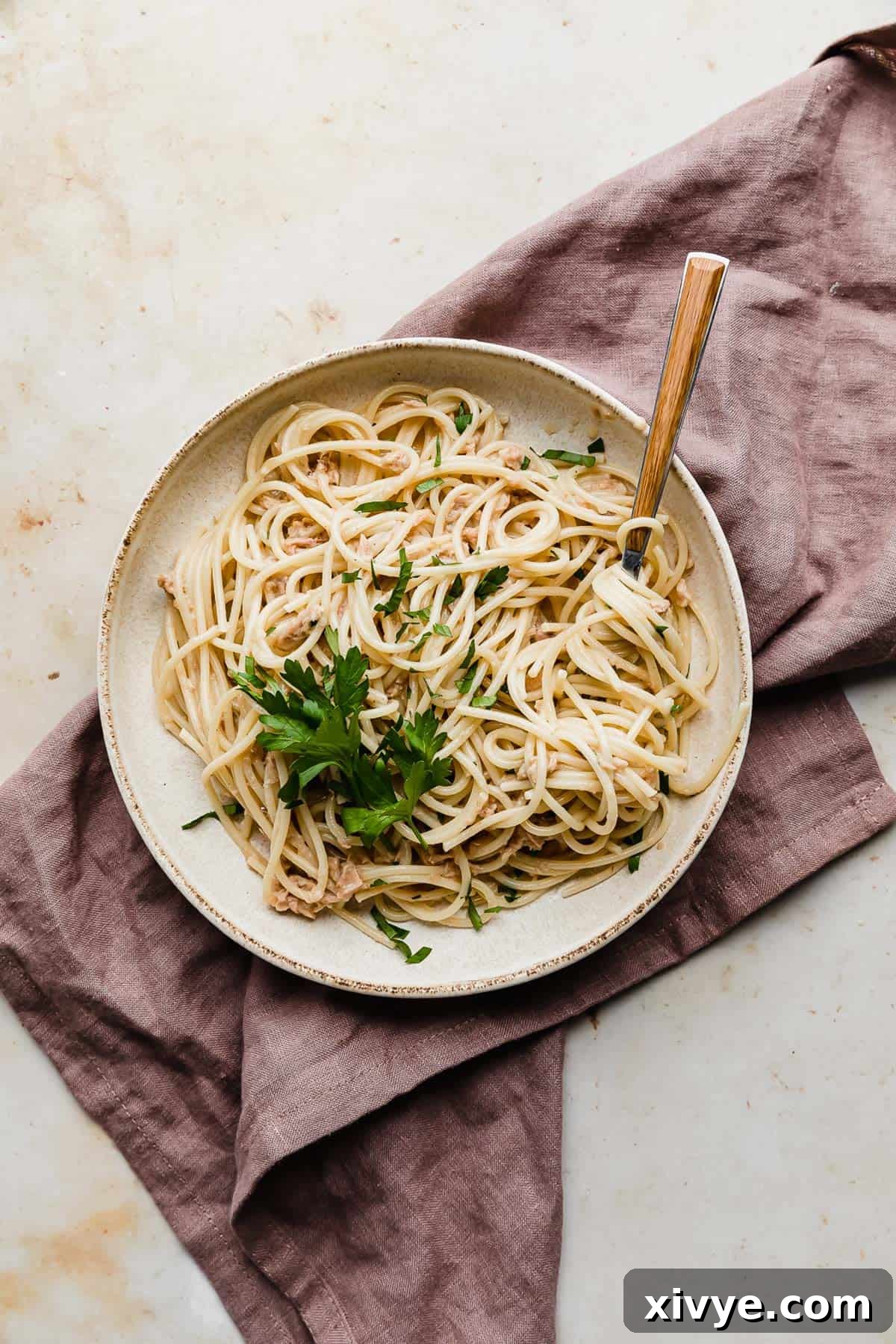 A white plate full of Tuna Carbonara topped with fresh parsley, on a lavender napkin.