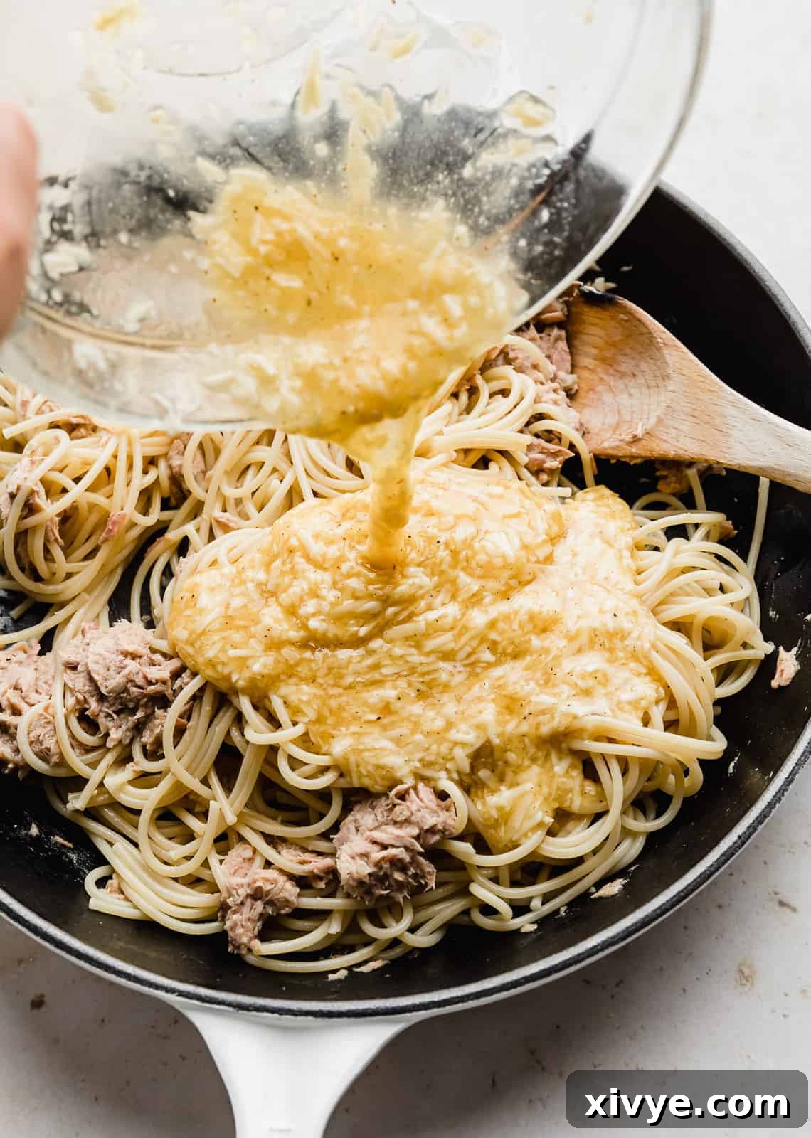 A yellow egg and cheese mixture being poured overtop of spaghetti noodles and tuna. 
