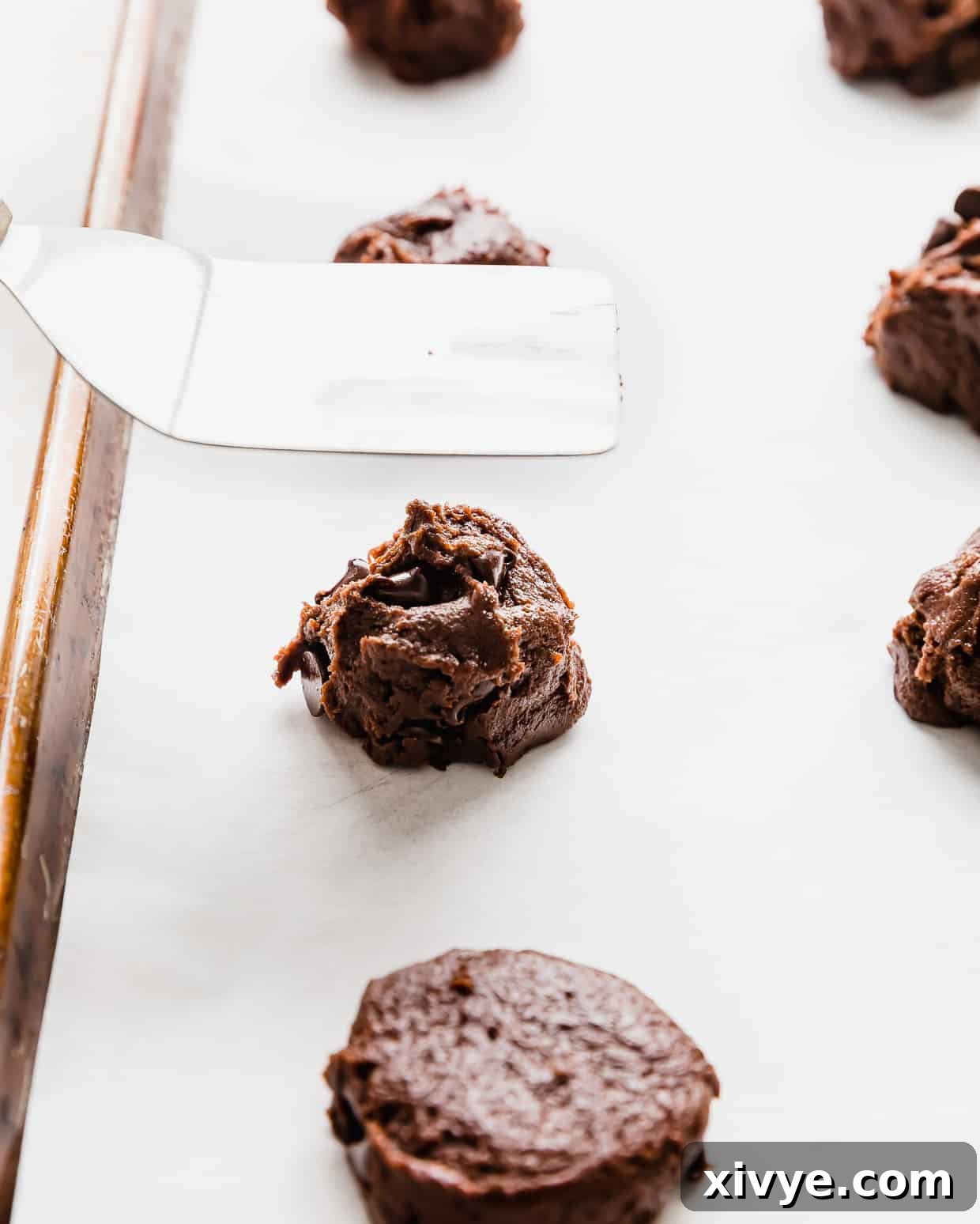 Cookie Ice Cream Delight 4 A metal flat head spatula hovering over a ball of double chocolate chip cookie dough on a baking sheet.