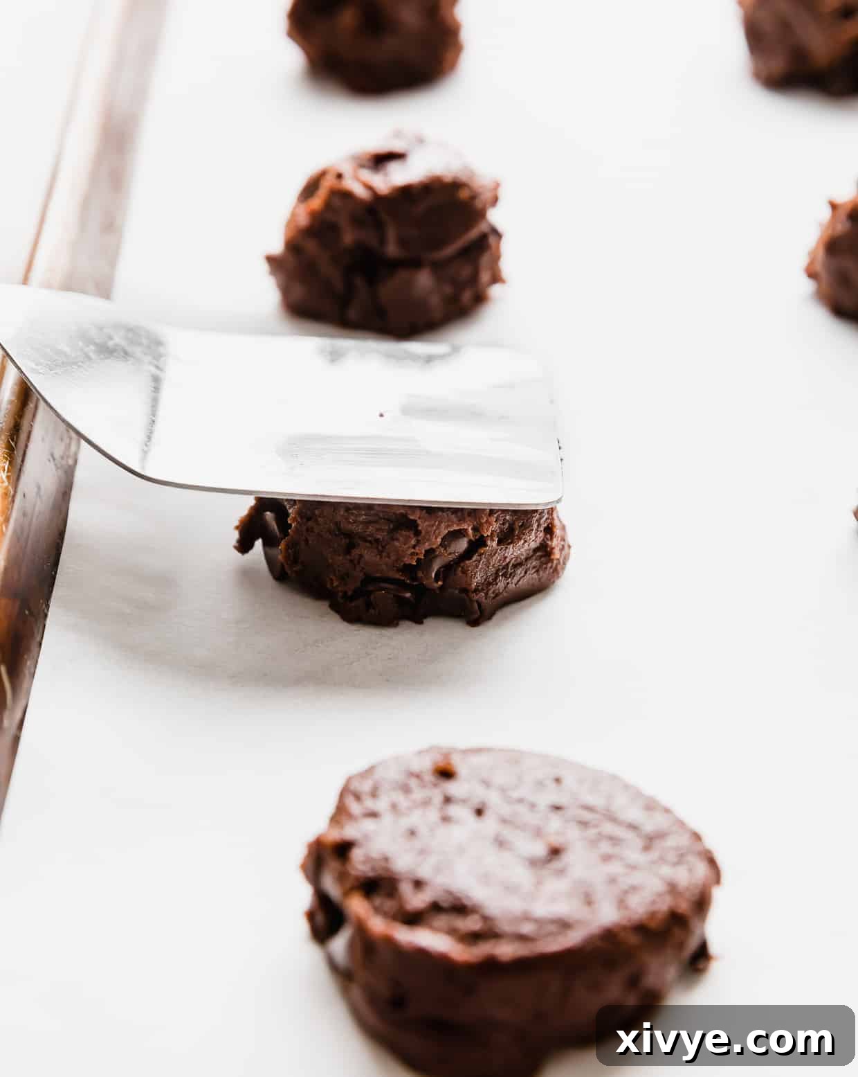 Cookie Ice Cream Delight 5 A metal spatula pressing down on a chocolate cookie dough ball on a baking sheet.