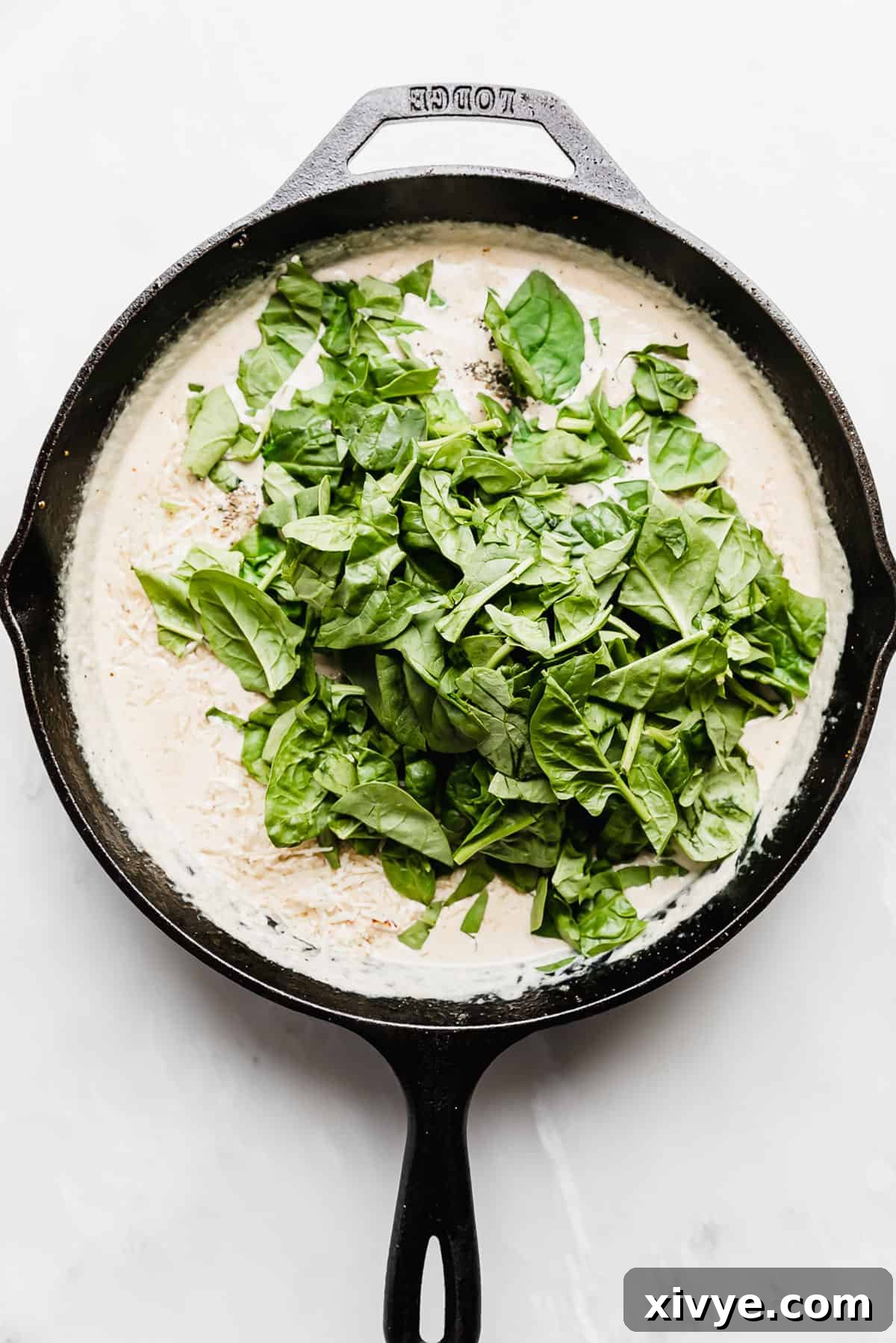 Spinach overtop a tan tuscan chicken gravy mixture in a skillet on a white background.