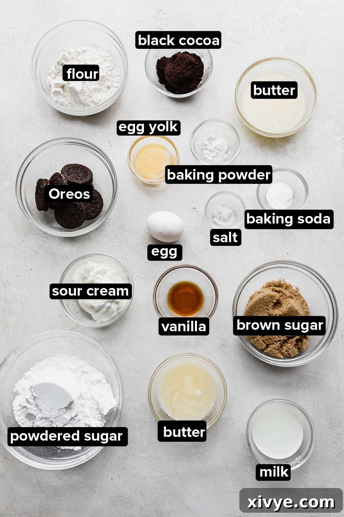 Oreo Donut ingredients, including flour, black cocoa powder, brown sugar, eggs, sour cream, and butter, neatly arranged in glass bowls on a gray background.