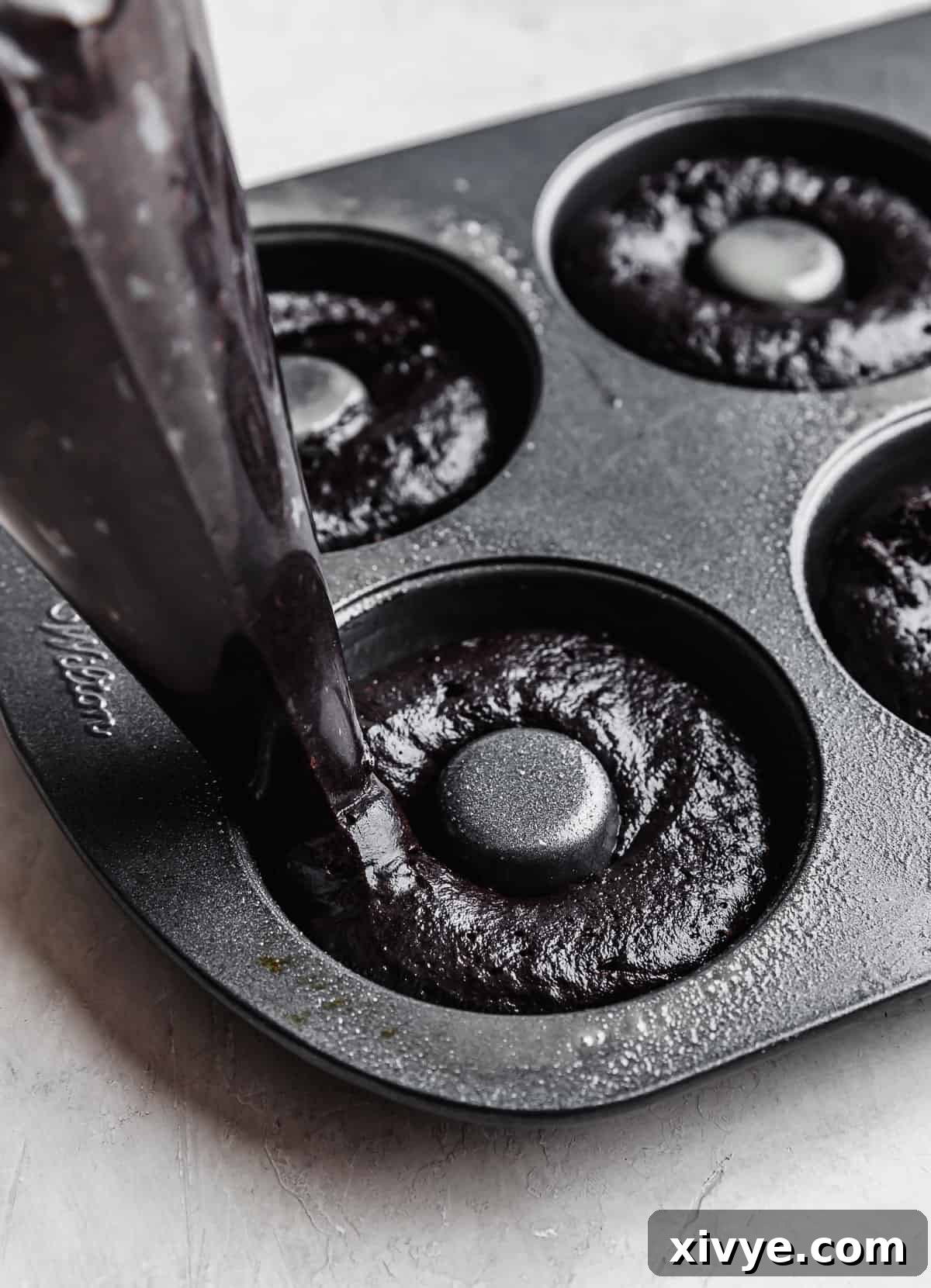A piping bag precisely dispensing Oreo Donut batter into the cavities of a donut pan.