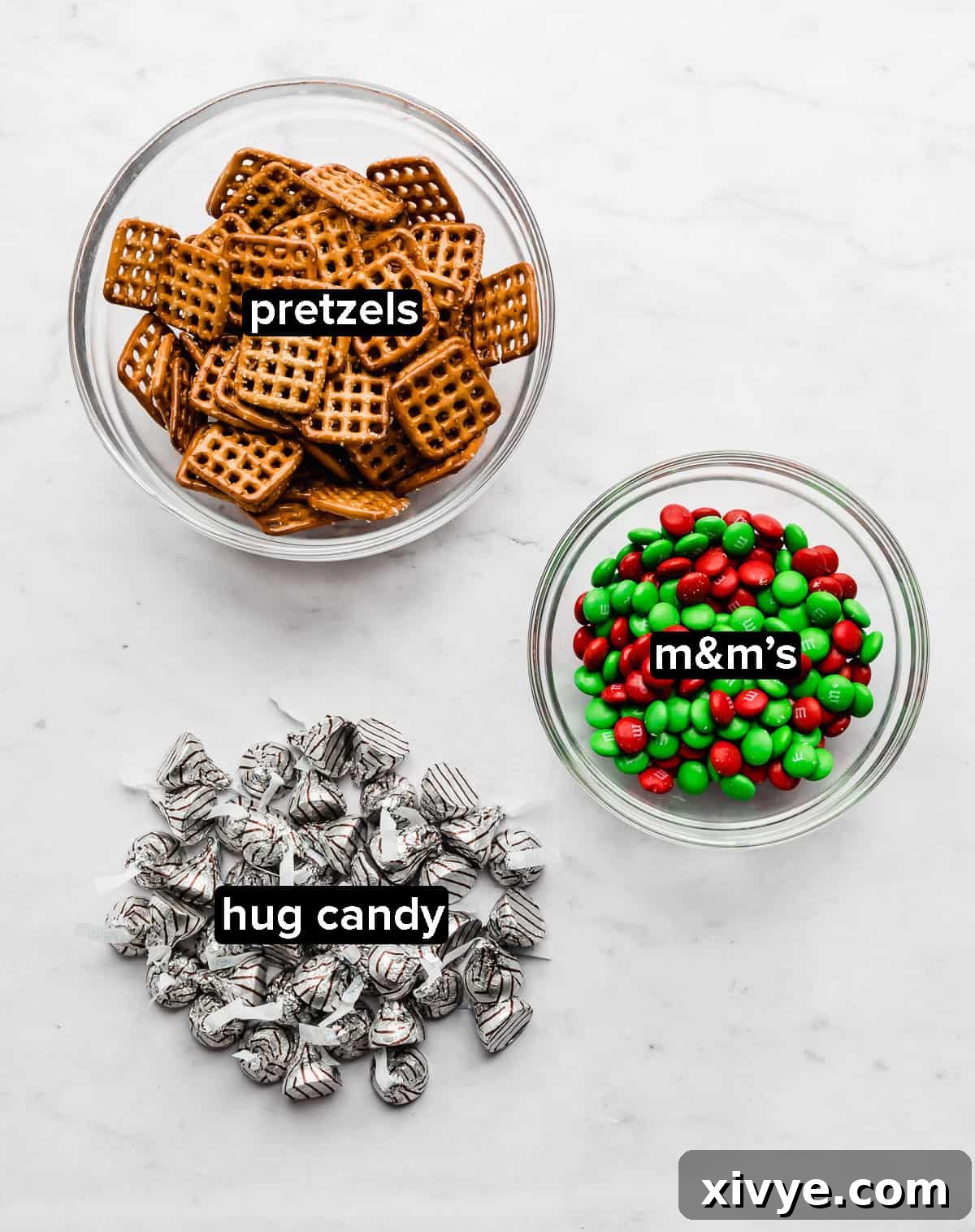 Yuletide Pretzel Bites 3 A colorful array of ingredients for Christmas Pretzel Hugs, including square pretzels, Hershey's Hugs, and festive red and green M&M's, arranged neatly on a elegant marble background.