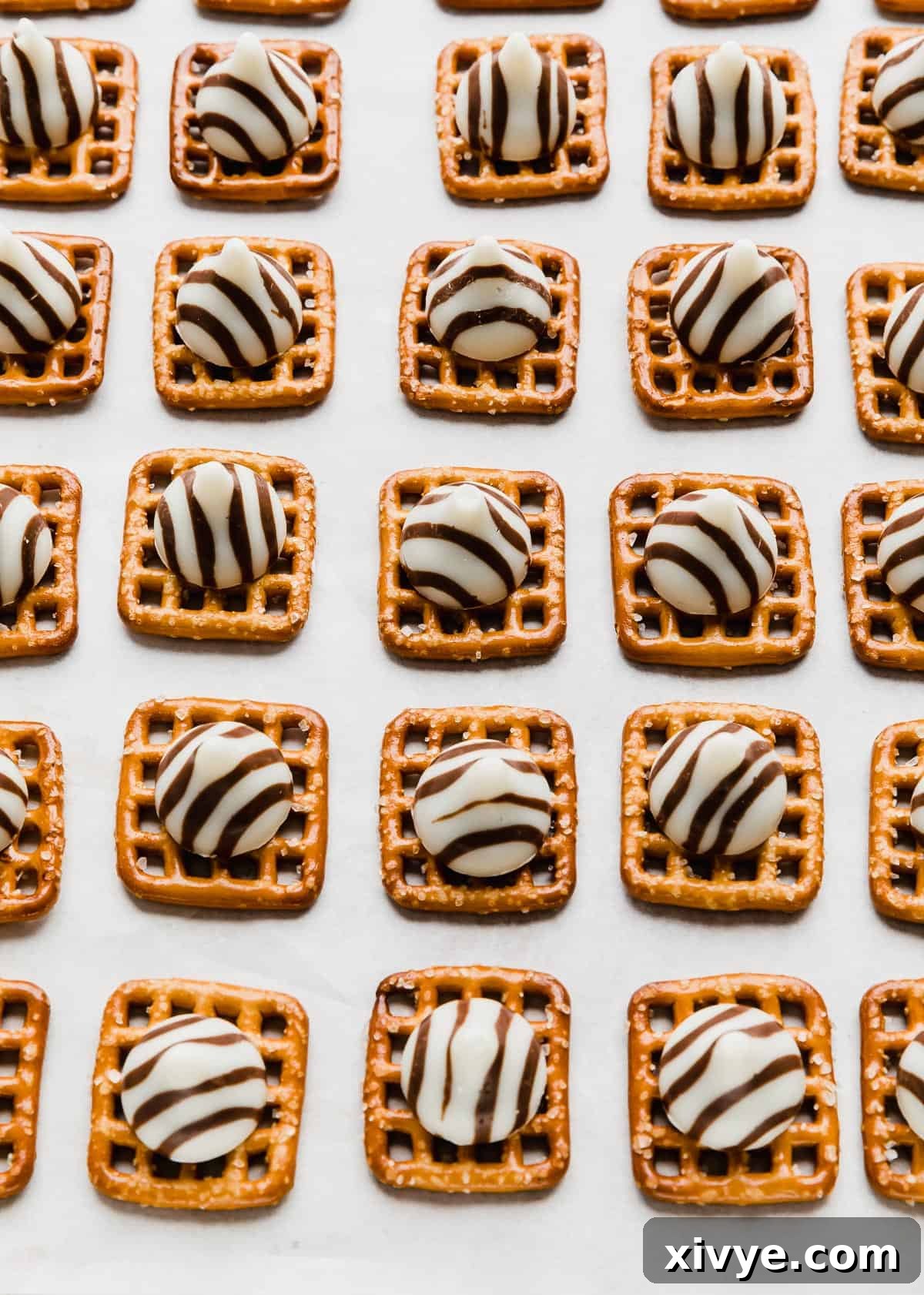 Yuletide Pretzel Bites 4 A baking sheet is filled with square pretzels, each meticulously topped with an unwrapped Hershey's Hug candy, ready for the oven.
