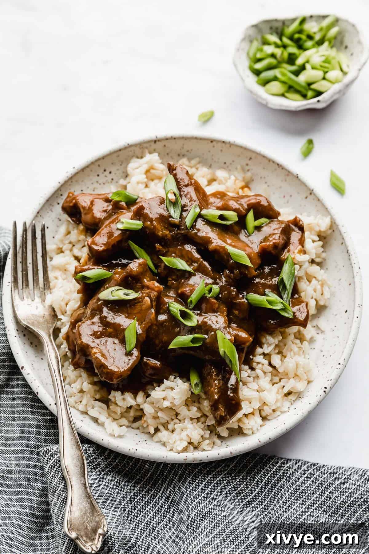 A white plate full of Slow Cooker Mongolian Beef on a bed of brown rice, garnished with green onions.