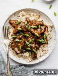A white plate with Slow Cooker Mongolian Beef topped with sliced green onions on brown rice.