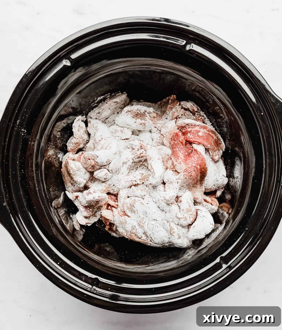 Flank steaks covered in cornstarch now placed in a black circular slow cooker, ready for the sauce ingredients.