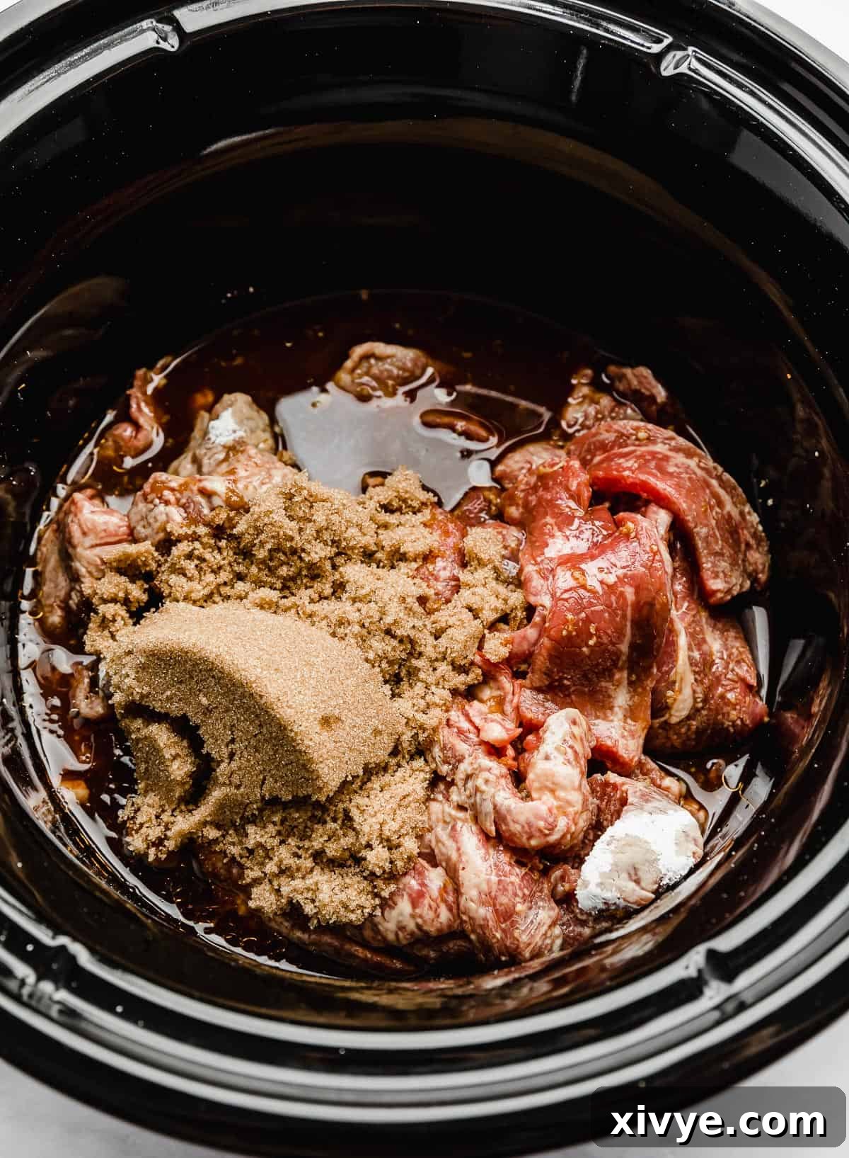 Brown sugar being poured over sliced flank steaks in a slow cooker with other sauce ingredients.