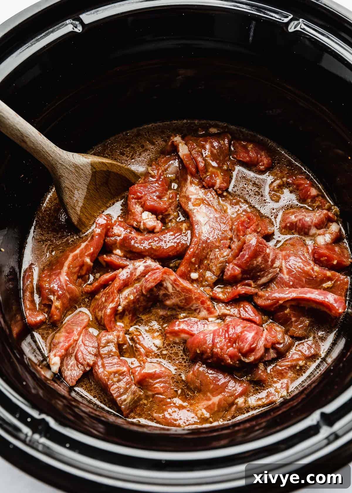 Raw flank steak strips in a slow cooker fully covered in the brown Mongolian beef sauce, ready for cooking.