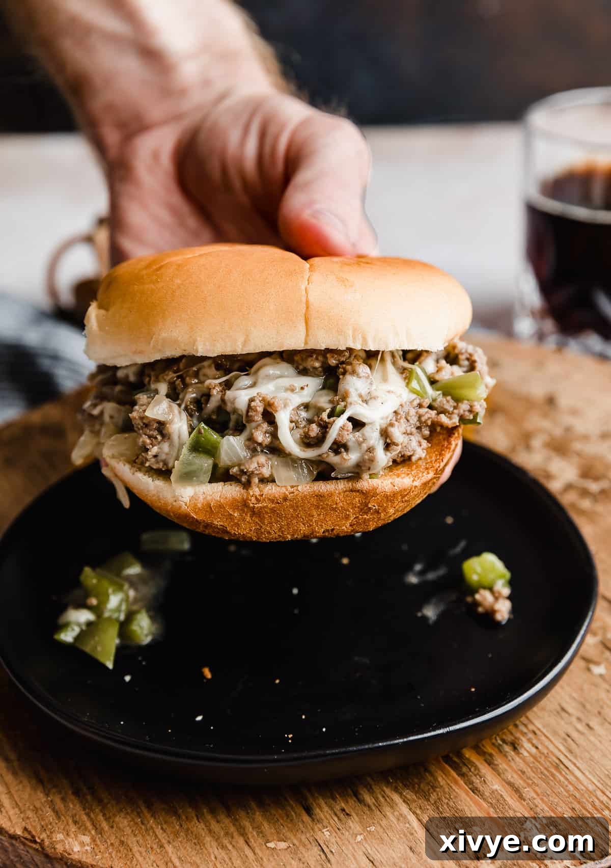 Philly Cheesesteak Joes 9 A hand gracefully holds a perfectly assembled Philly Cheesesteak Sloppy Joe on a toasted bun, poised over a black plate, ready to be enjoyed.
