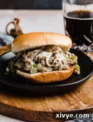 A Philly Cheesesteak Sloppy Joe on a black plate with a clear glass full of brown liquid in the background.