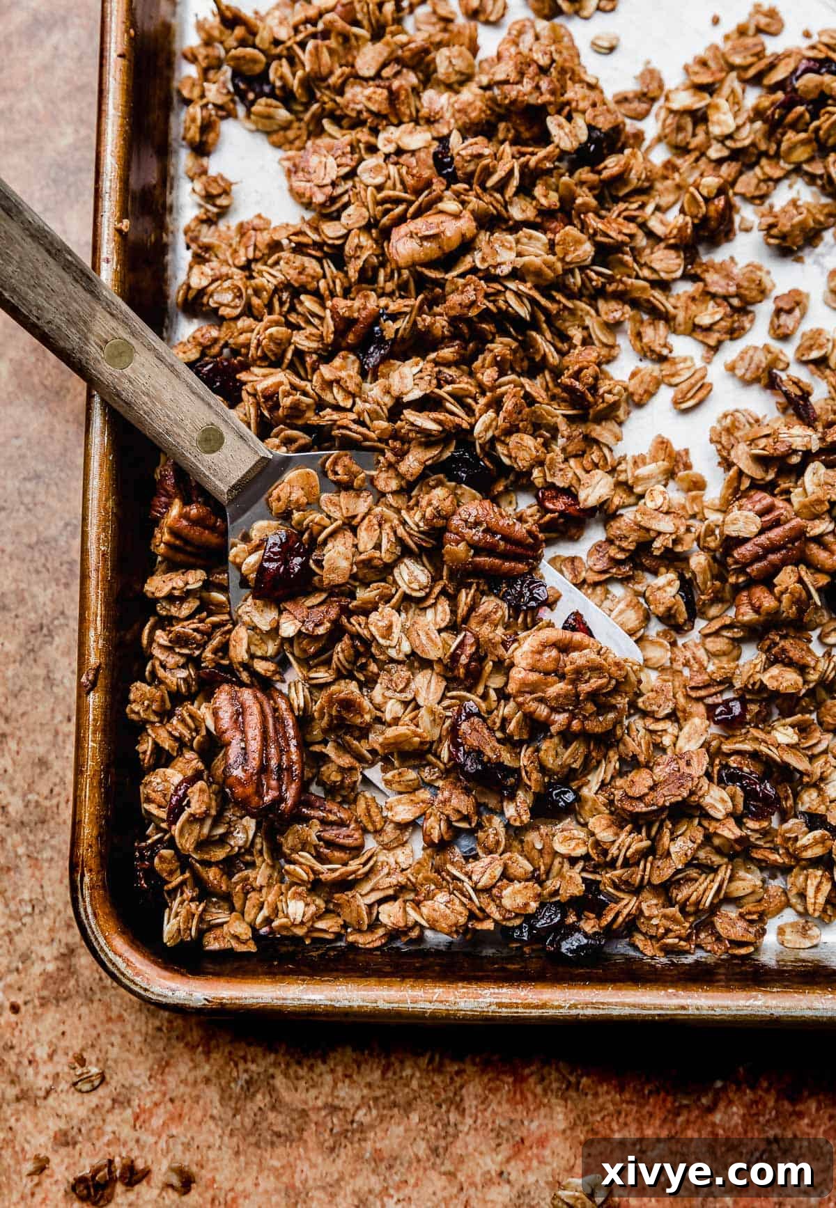 Festive Gingerbread Granola 12 A metal spatula carefully scooping up deep amber-colored gingerbread granola from a baking sheet, showing off its clusters.