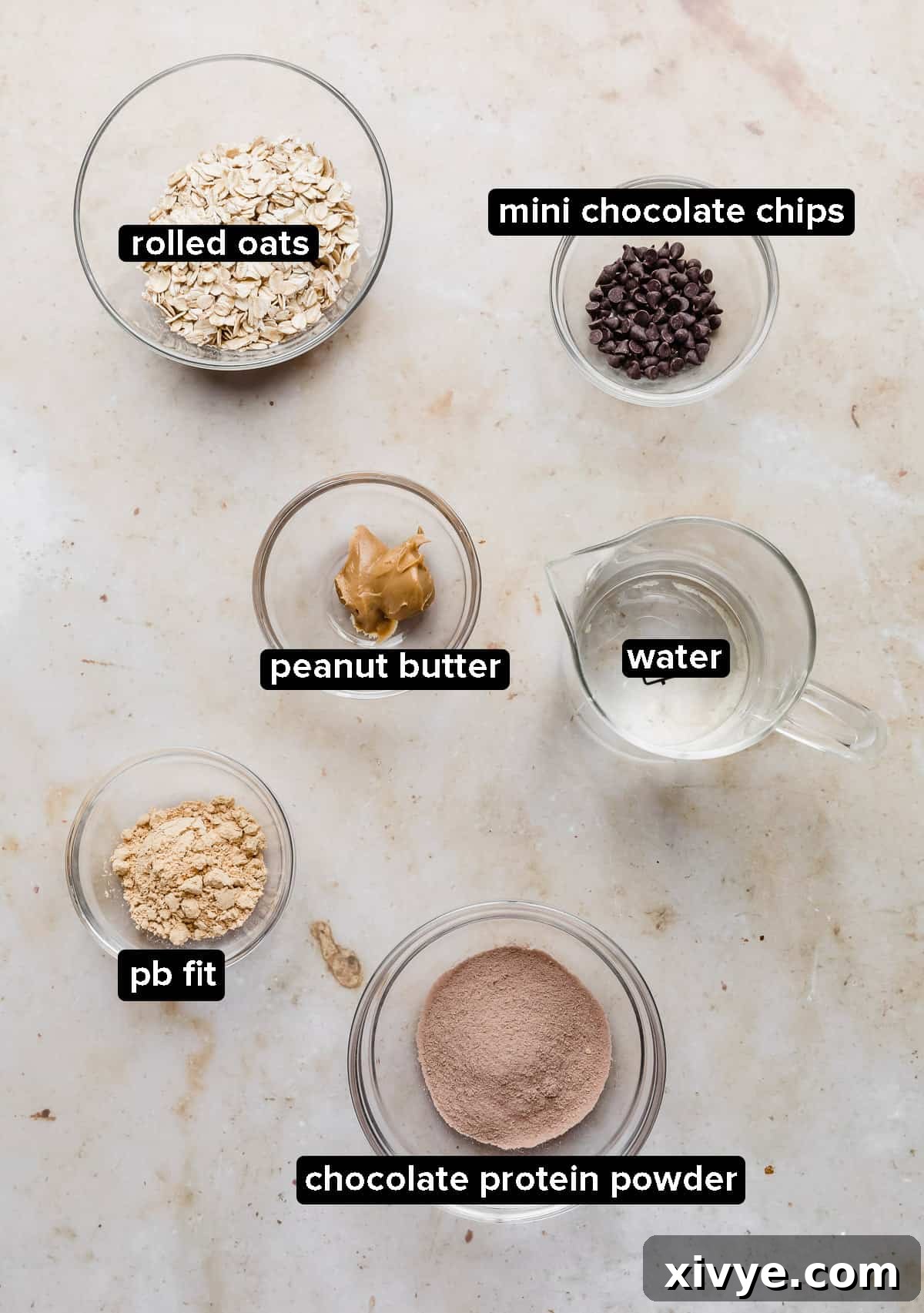 Chocolate Peanut Butter Protein Oatmeal ingredients in glass bowls on a tan marble background.