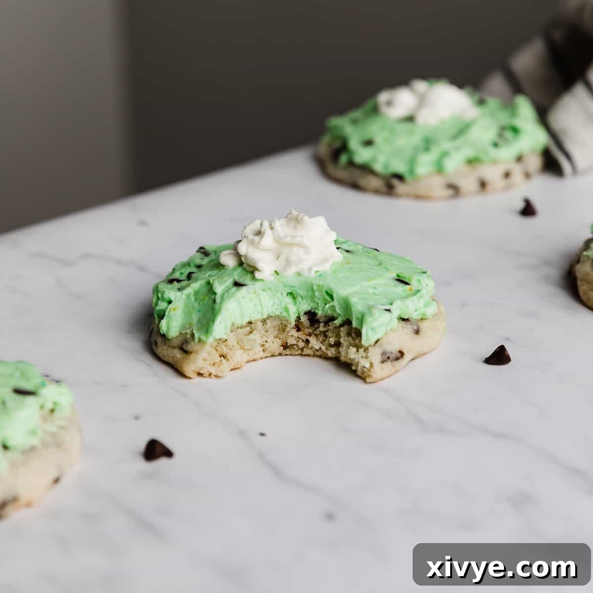 A delectable copycat Crumbl Mint Chip Ice Cream Cookie with a bite taken out, featuring a vibrant mint green frosting, mini chocolate chips, and a dollop of whipped cream, for a refreshing treat.