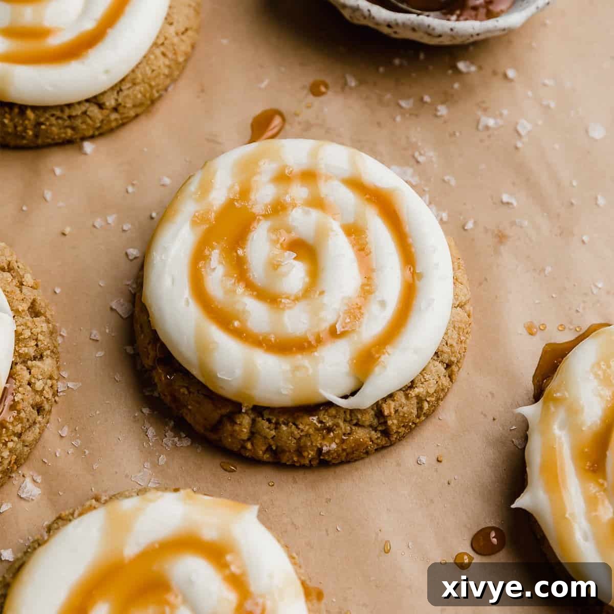 A luxurious Salted Caramel Cheesecake Cookie, boasting a graham cracker base, topped with creamy white frosting, a swirl of caramel, and a sprinkle of flaky salt, presented on tan parchment paper.