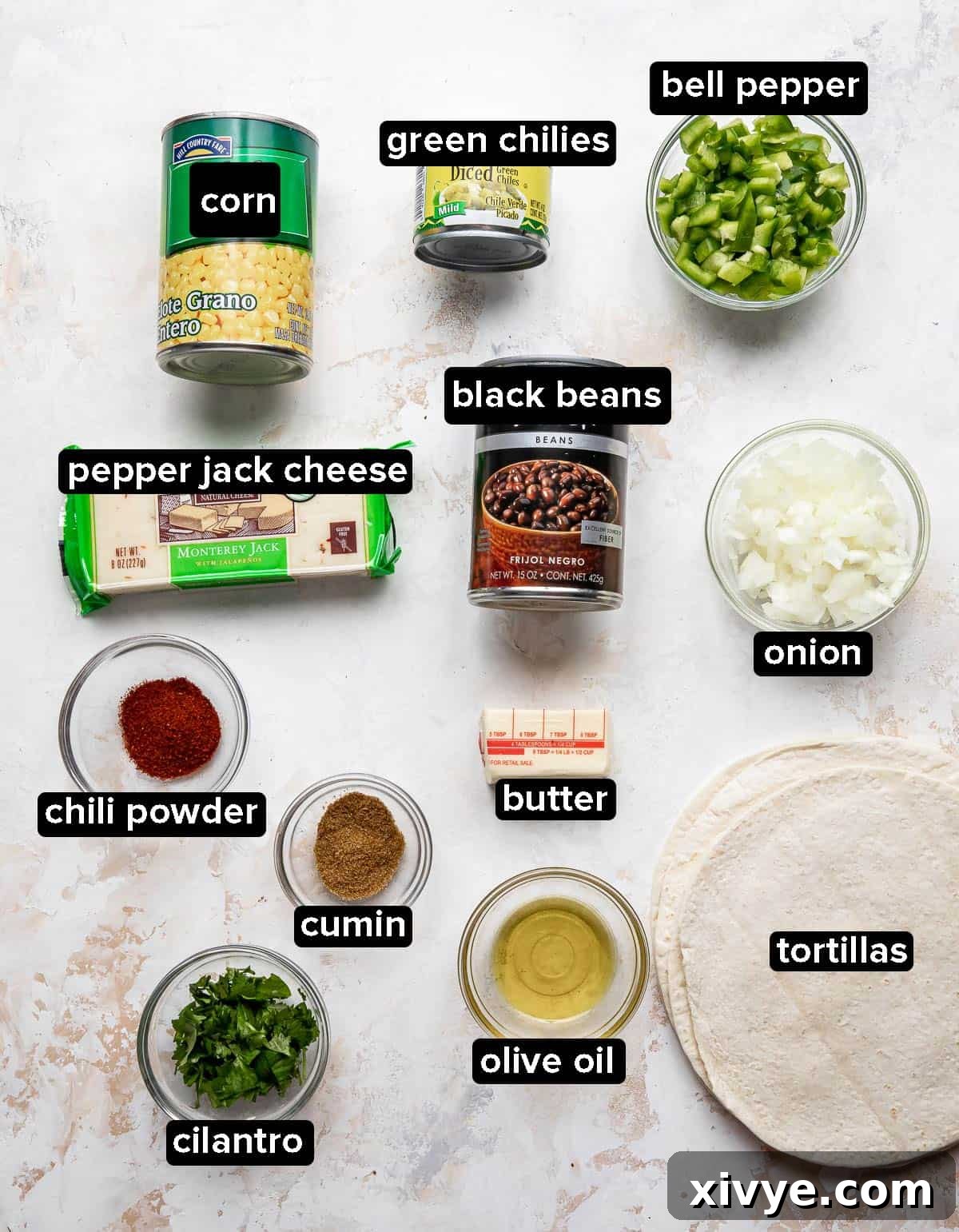 A vibrant flat lay of Black Bean and Corn Quesadilla ingredients including flour tortillas, canned corn, black beans, diced green chiles, fresh green bell pepper and onion, and pepper jack cheese, arranged on a white textured background.