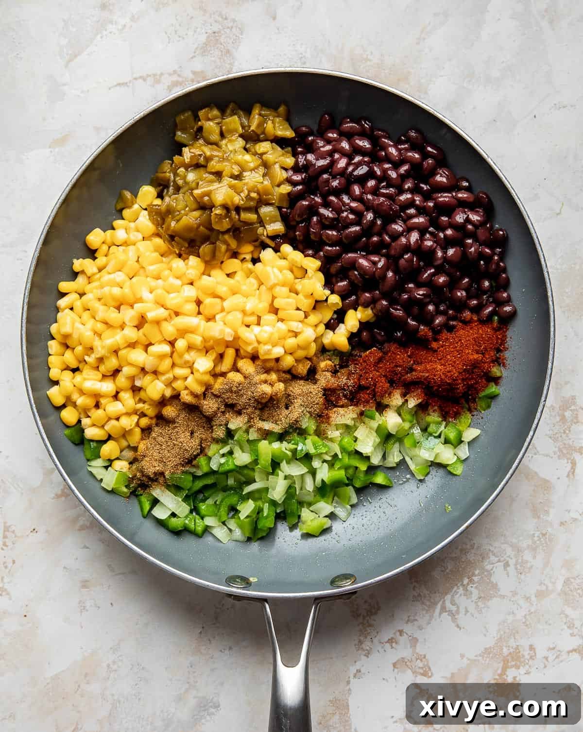A gray skillet with vibrant corn, green bell pepper, black beans, chili powder, and diced green chilies, beginning to warm through and combine their flavors.