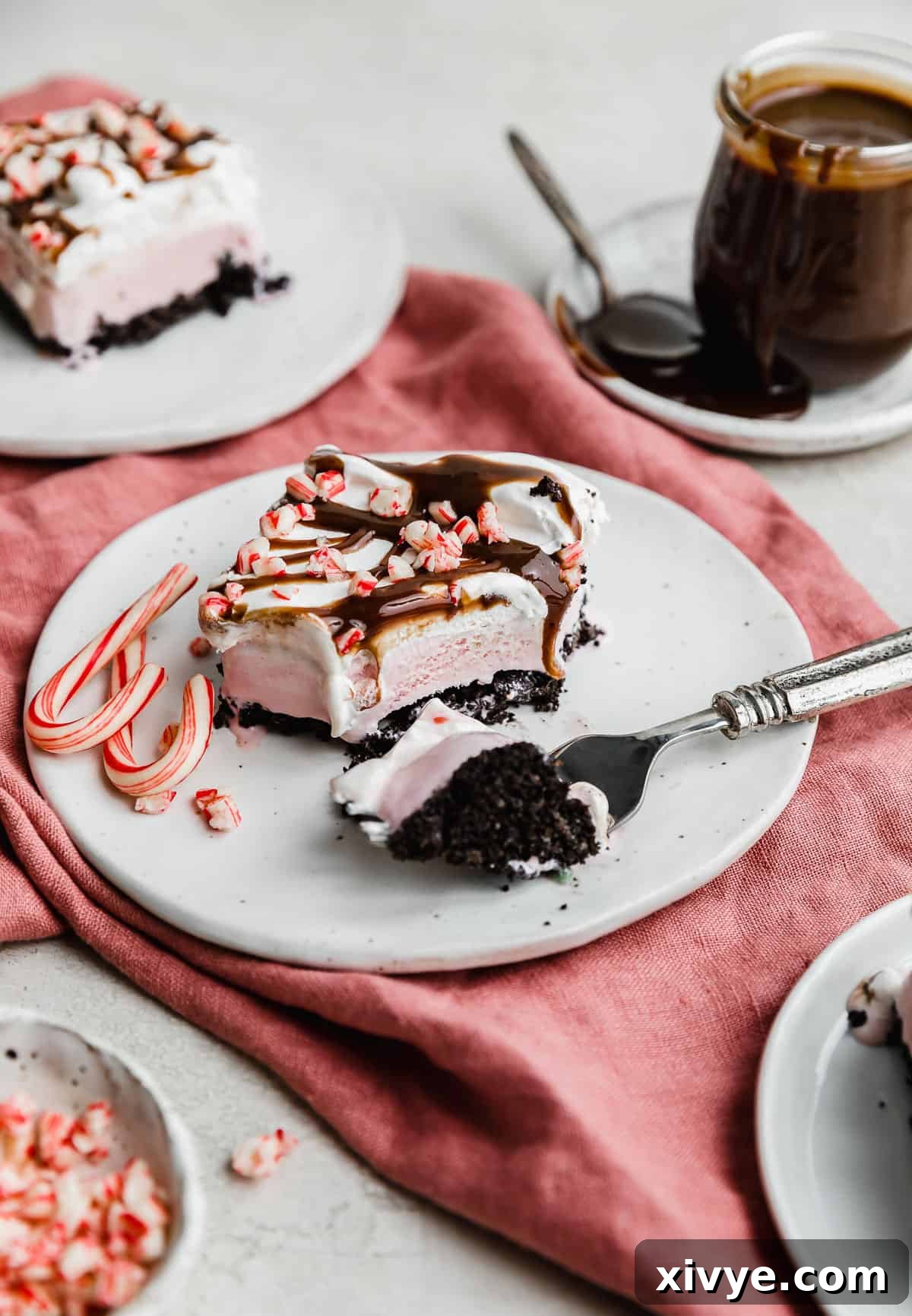 A perfectly sliced Christmas Ice Cream Cake on a white plate, showcasing its distinct layers of dark Oreo crust, light pink peppermint ice cream, fluffy white whipped cream, and a sprinkle of crushed candy canes.