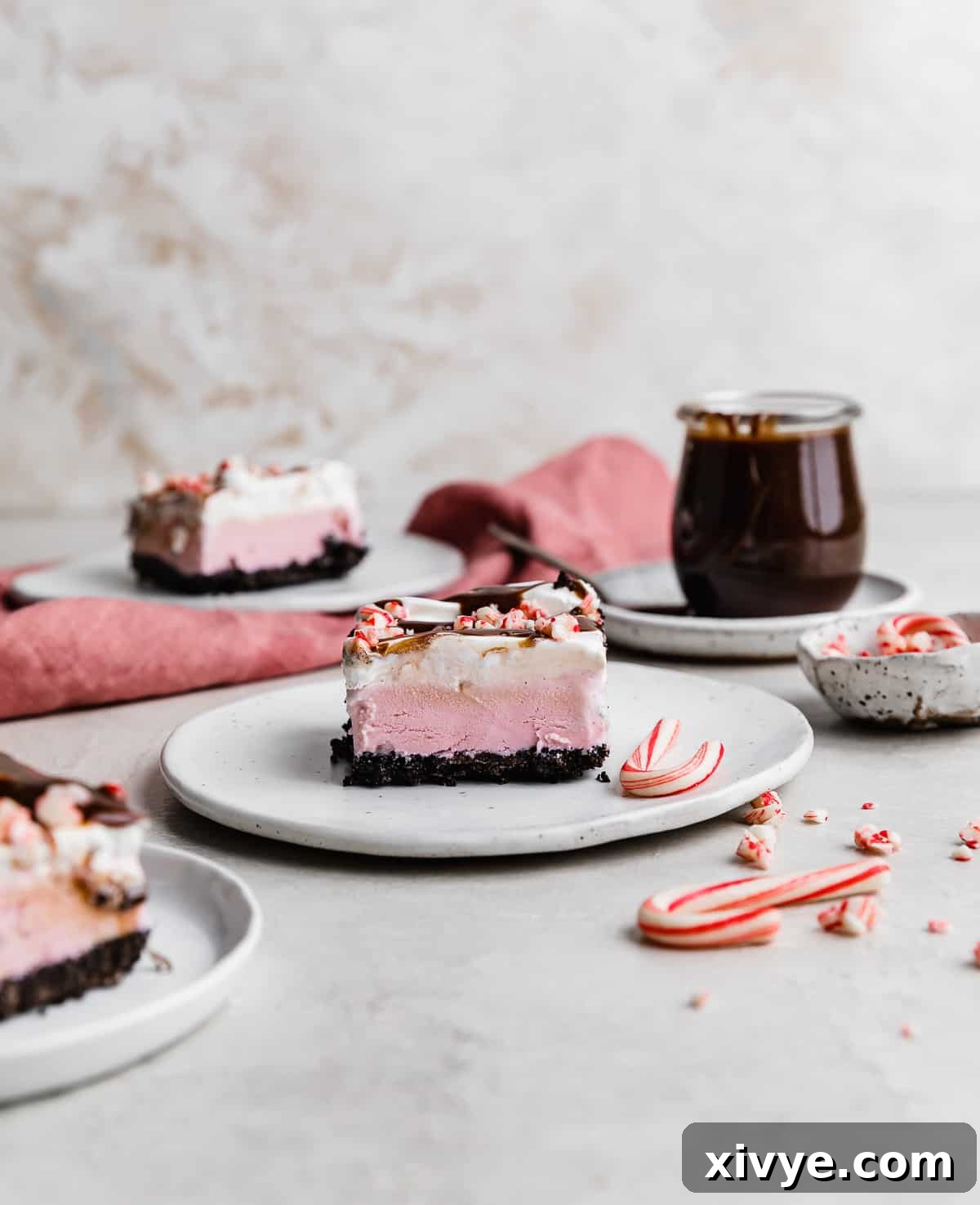 A delectable slice of Christmas Ice Cream Cake on a white plate, with a jar of rich hot fudge subtly blurred in the background, hinting at the indulgent toppings.