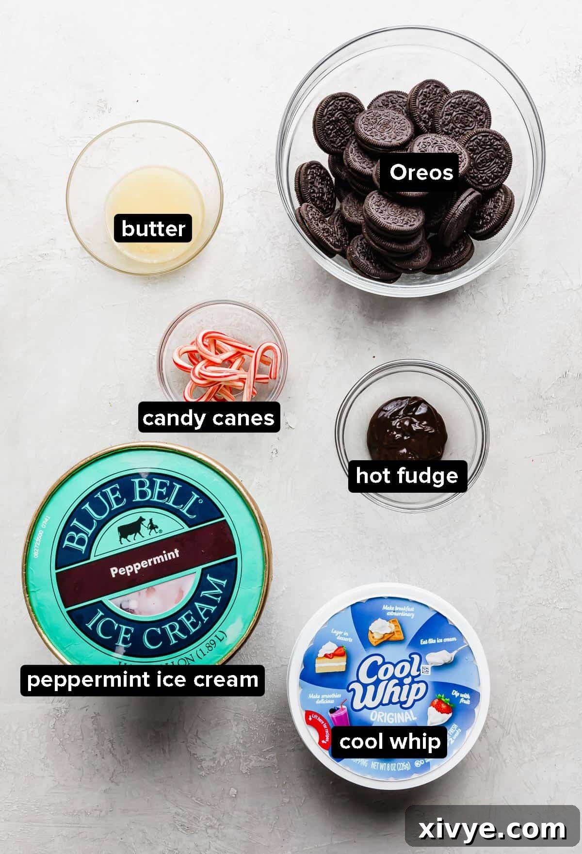 All the core ingredients for making Christmas Ice Cream Cake laid out on a light gray background: a package of Oreos, a stick of butter, a container of peppermint ice cream, Cool Whip, and crushed candy canes.