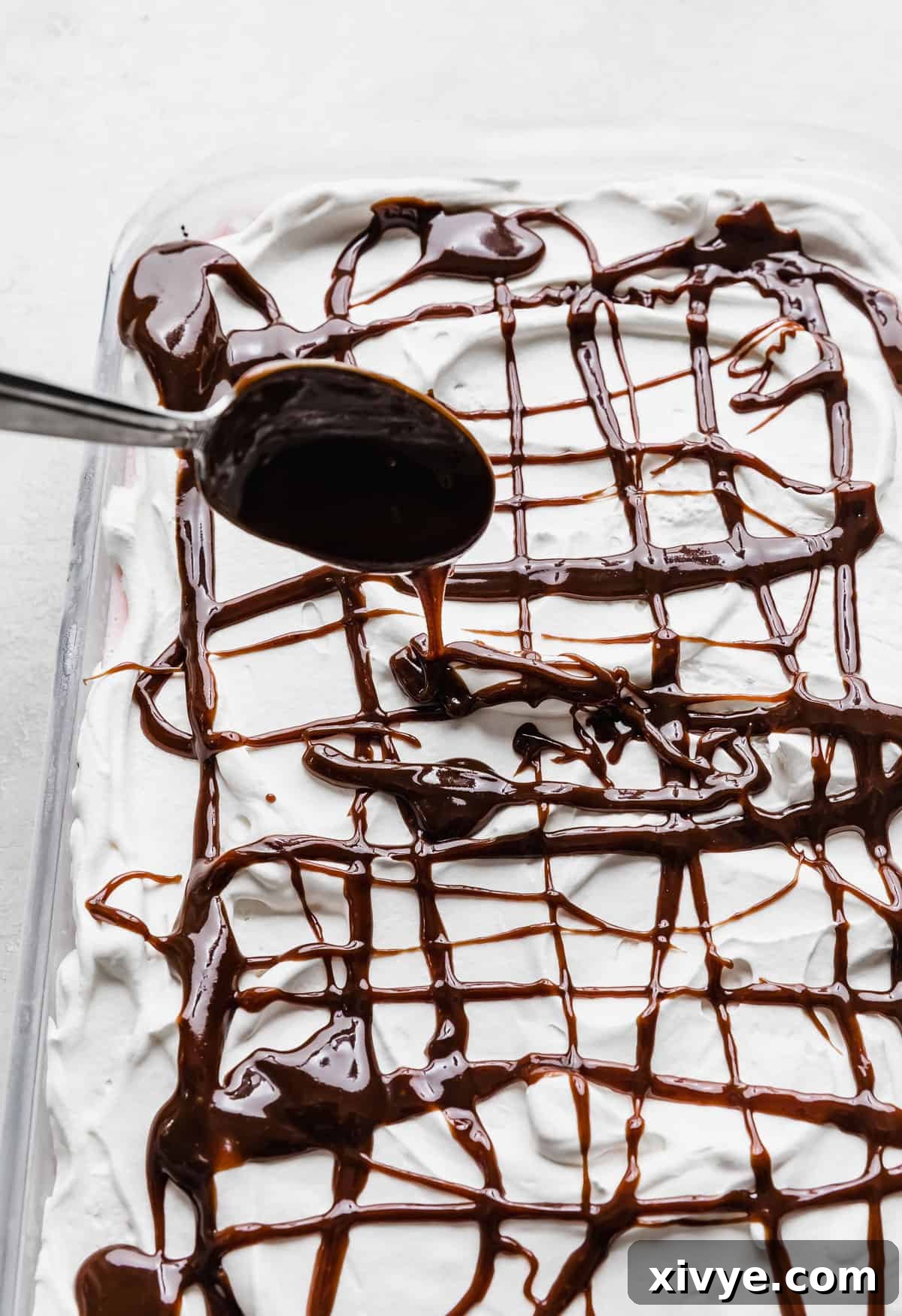 A spoon expertly drizzling homemade hot fudge sauce in a luscious pattern over the white whipped cream topping of the peppermint Christmas ice cream cake.