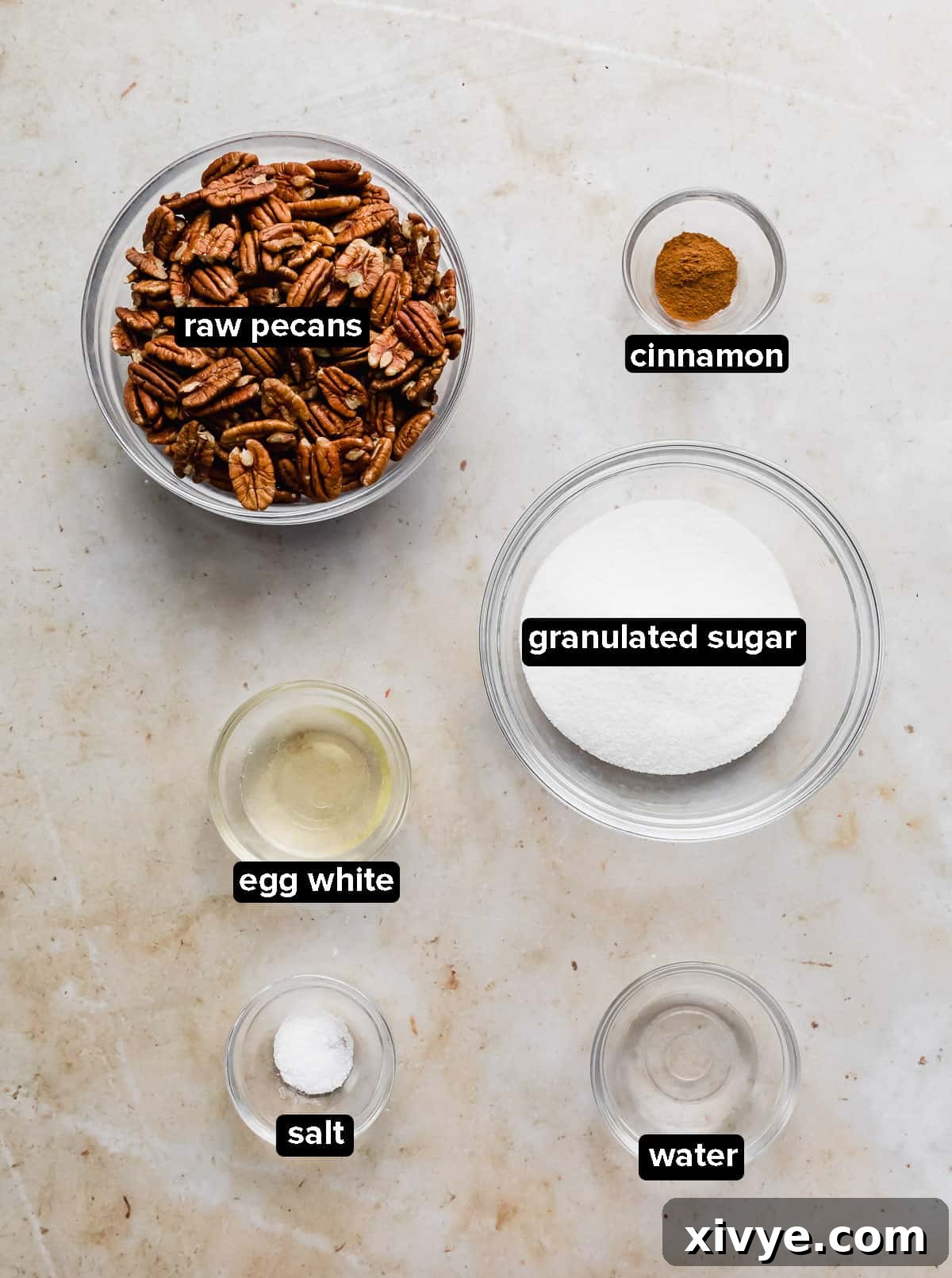 Sweet Glazed Pecans 3 Assorted ingredients for candied pecans, including egg white, sugar, cinnamon, and raw pecans, displayed in glass bowls on a marble background.