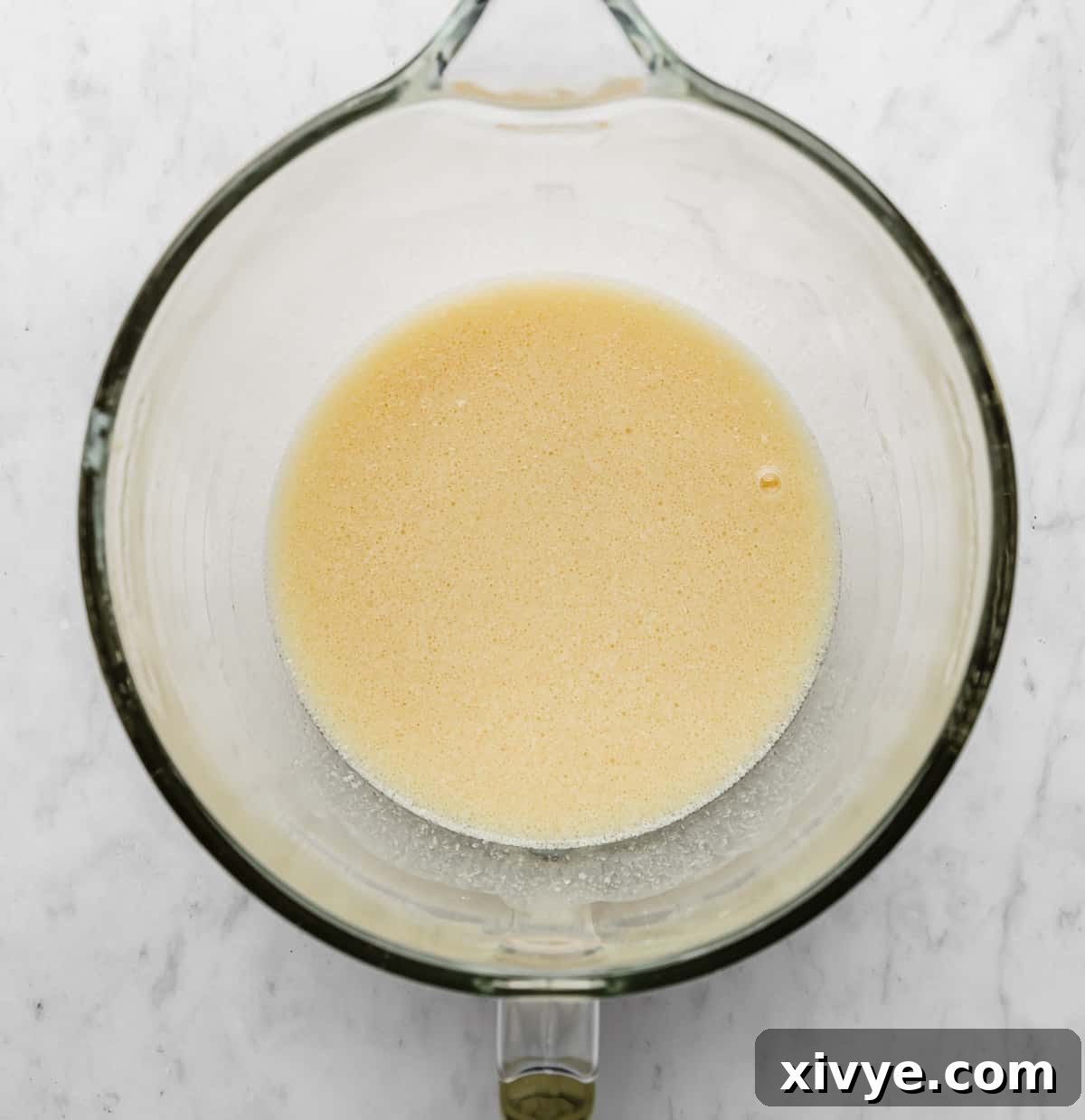 A light yellow colored liquid mixture in a glass mixing bowl on a white marble background.