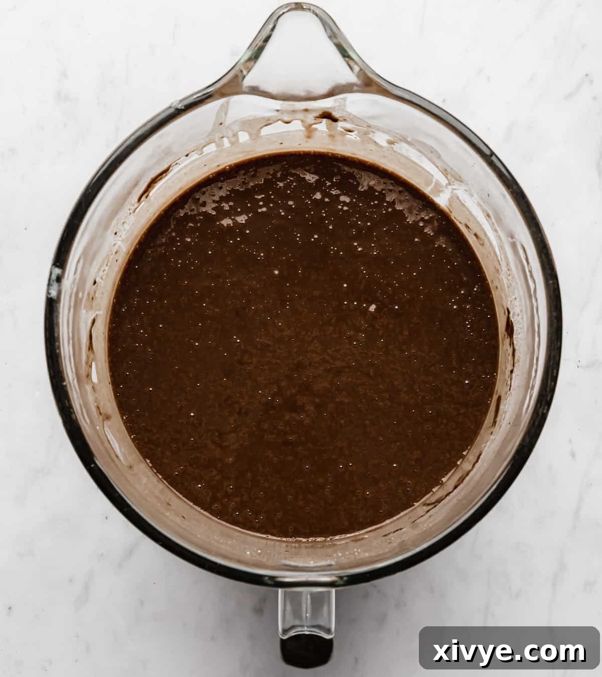 Chocolate Bundt Cake batter in a glass stand mixer bowl on a white marble background.