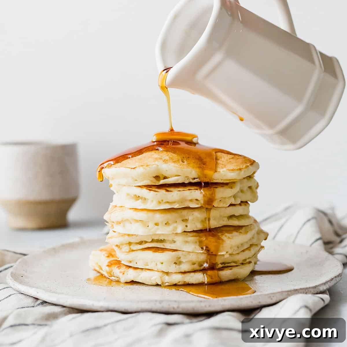 Maple syrup being poured overtop a stack of fluffy buttermilk pancakes on a white plate.