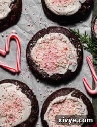 Peppermint Bark Cookies topped with white chocolate and crushed candy canes, on a white parchment paper that's on a forest green background.