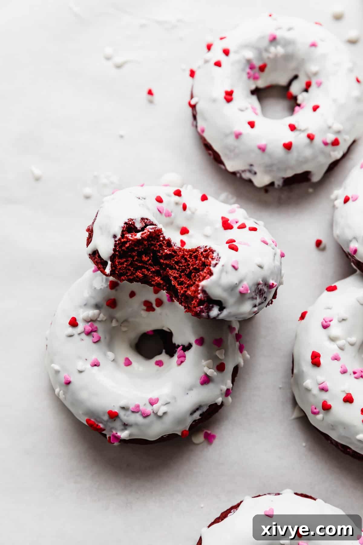 Velvet Dream Doughnuts 2 Red Velvet Donuts topped with a pristine white chocolate glaze and festive heart sprinkles, presented on a clean white background.