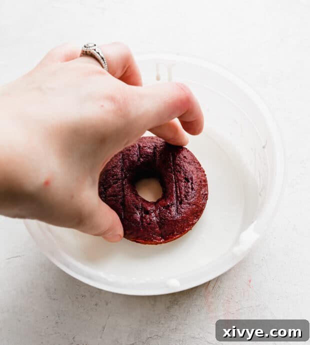 Velvet Dream Doughnuts 8 A hand gently dipping a vibrant red velvet donut into a bowl of creamy white chocolate glaze, creating a perfect coating.