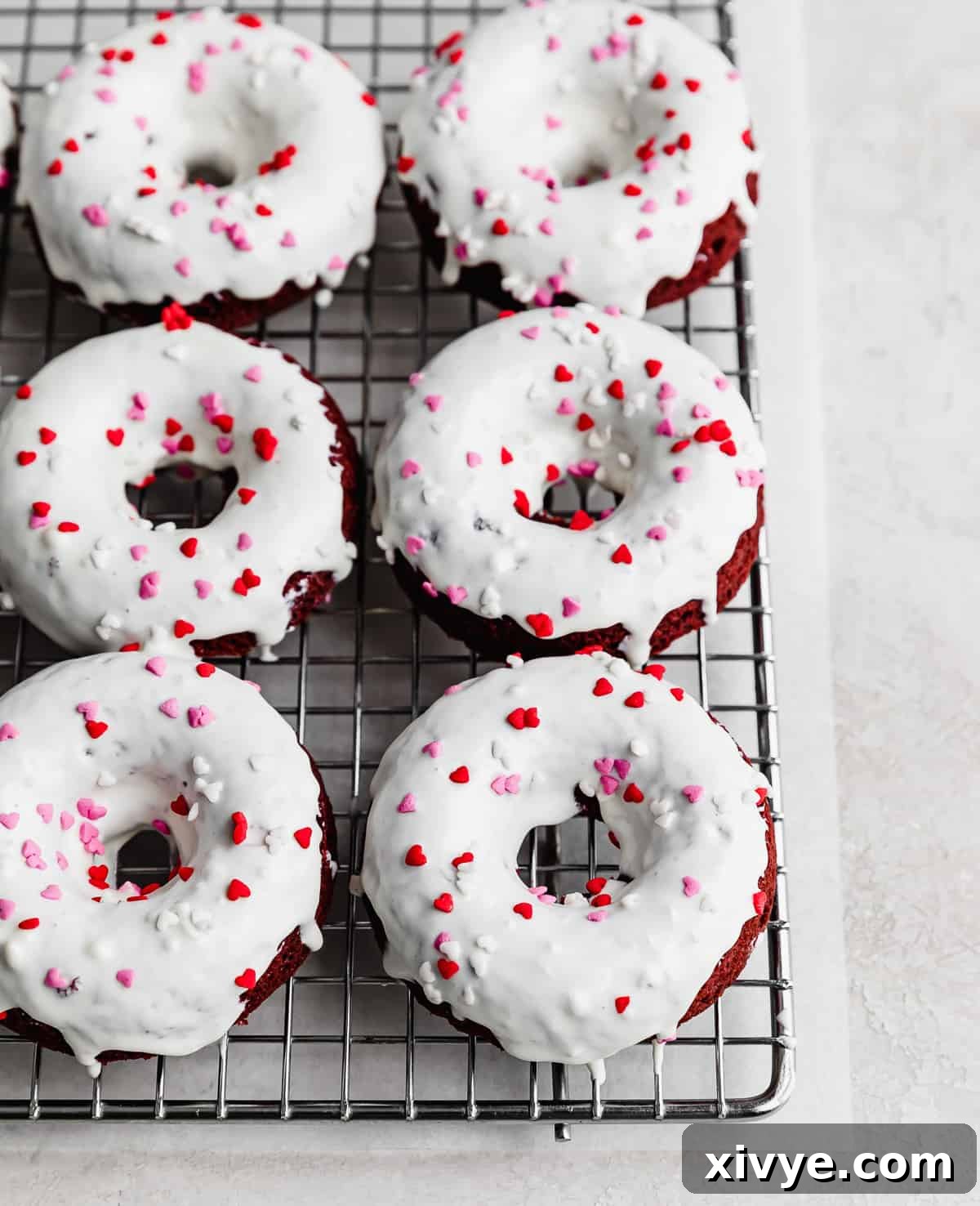 Velvet Dream Doughnuts 10 A cooling rack adorned with freshly baked Red Velvet Donuts, elegantly coated in a glistening white chocolate glaze and sprinkled with heart-shaped decorations.