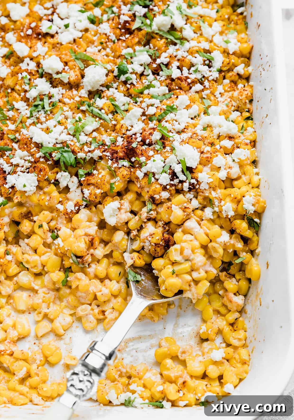 A large serving spoon elegantly scoops a portion of the creamy and vibrant Mexican Street Corn Casserole from a white baking dish, ready to be served.