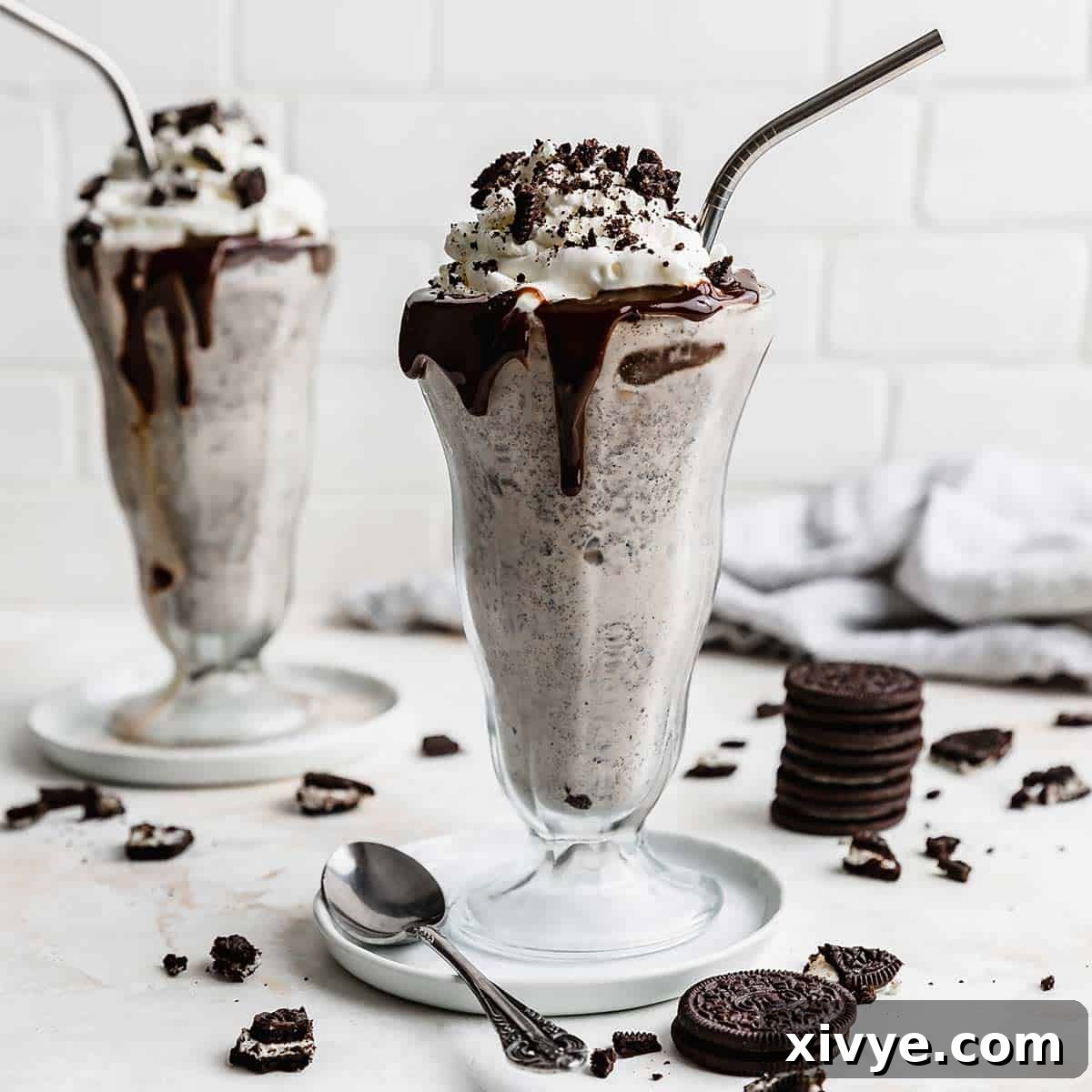 Irresistible Oreo Creations 3 Two milkshake glasses filled with the best Oreo Milkshake recipe, against a white brick background with Oreo crumbs scattered around the cups.