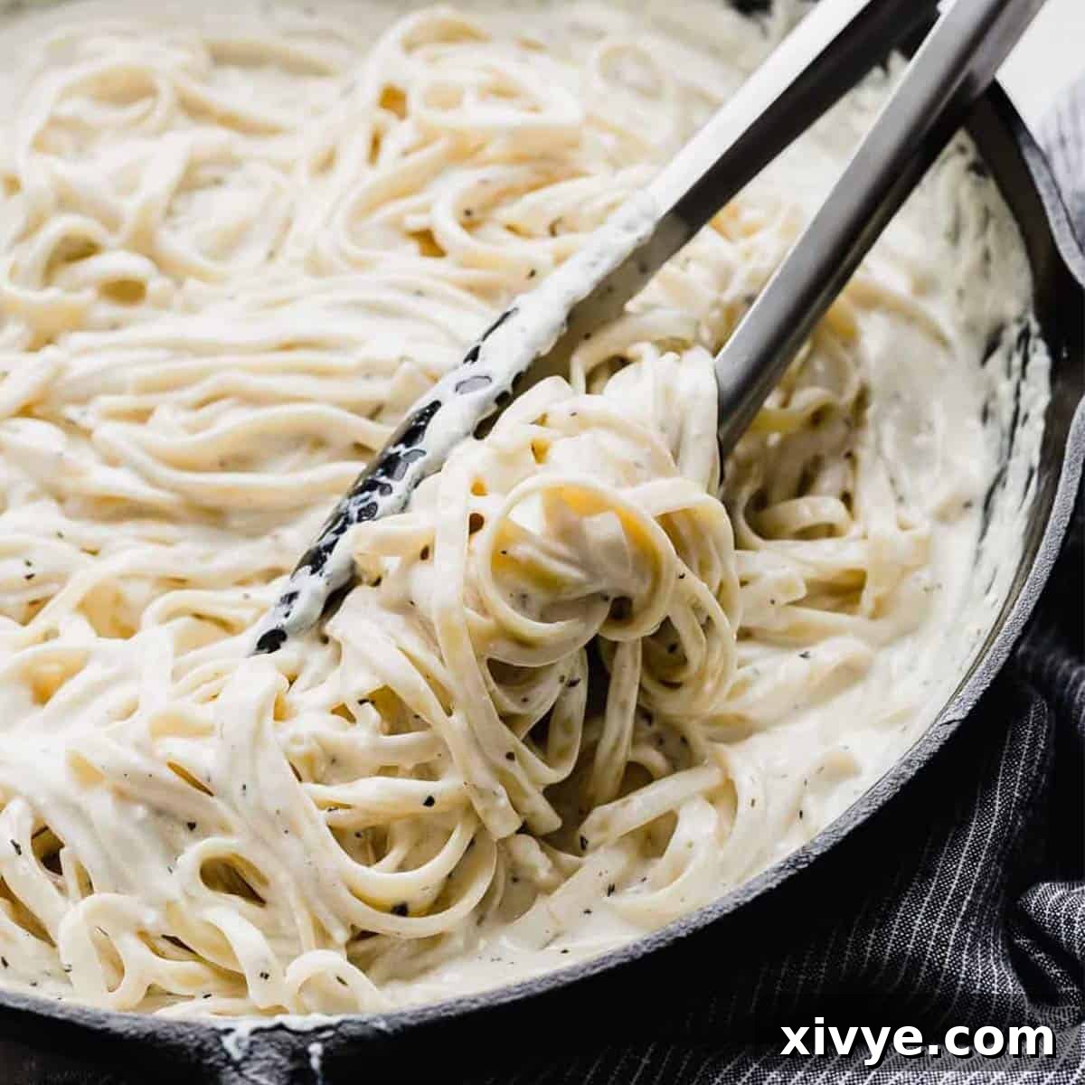 A black skillet filled with creamy Fettuccine Alfredo, with kitchen tongs expertly mixing the rich, flavorful pasta to ensure every strand is coated.