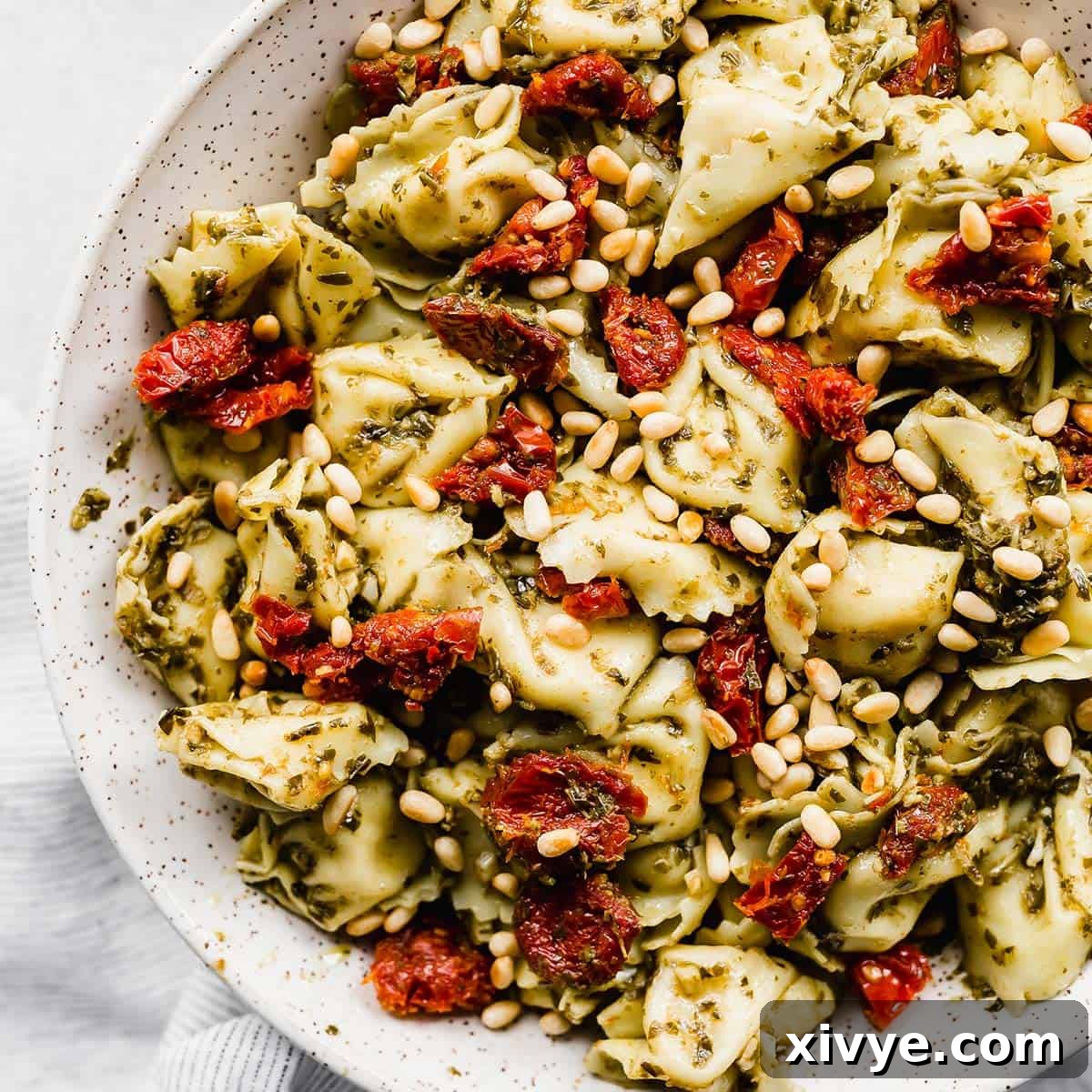 A close-up photo of Pesto Tortellini Pasta Salad, beautifully tossed with sun-dried tomatoes and pine nuts, presented in an elegant white bowl.