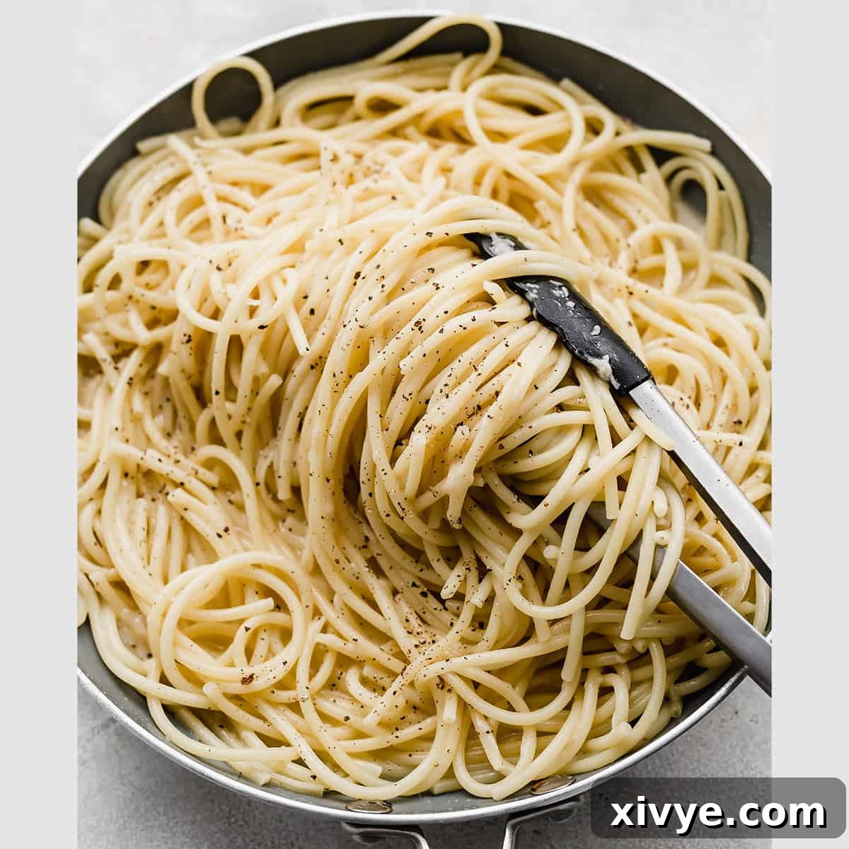 Thick Bucatini Cacio e Pepe noodles swirling in a skillet pan, with kitchen tongs gently turning the pasta to coat it evenly.