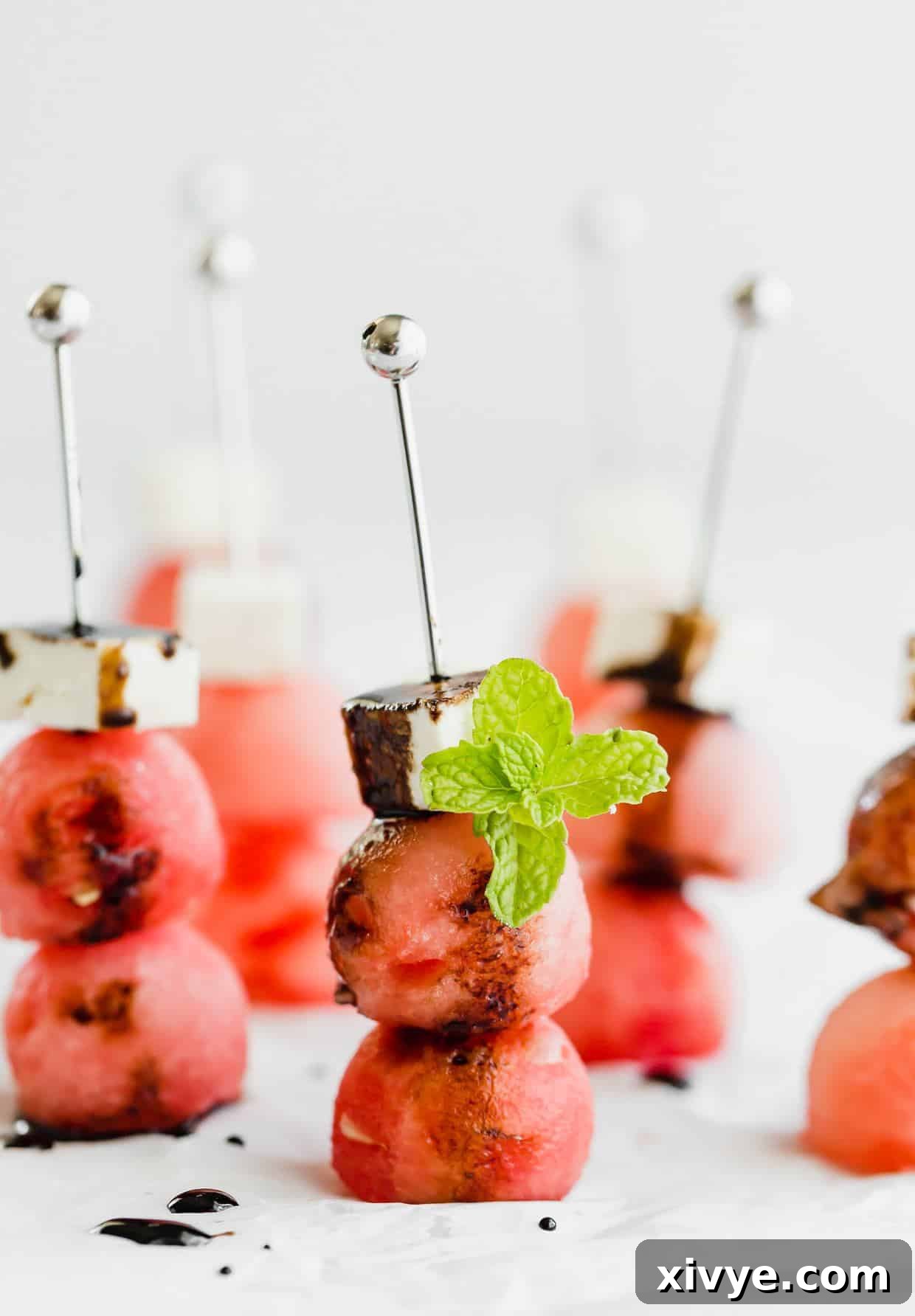 Two watermelon balls and a block of feta on small skewers, drizzled with balsamic reduction and a mint leaf for garnish.