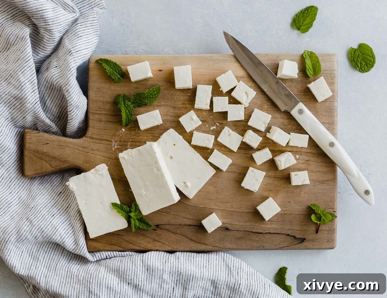 Overhead view of a wooden cutting board with a block of feta cheese cut into small squares.