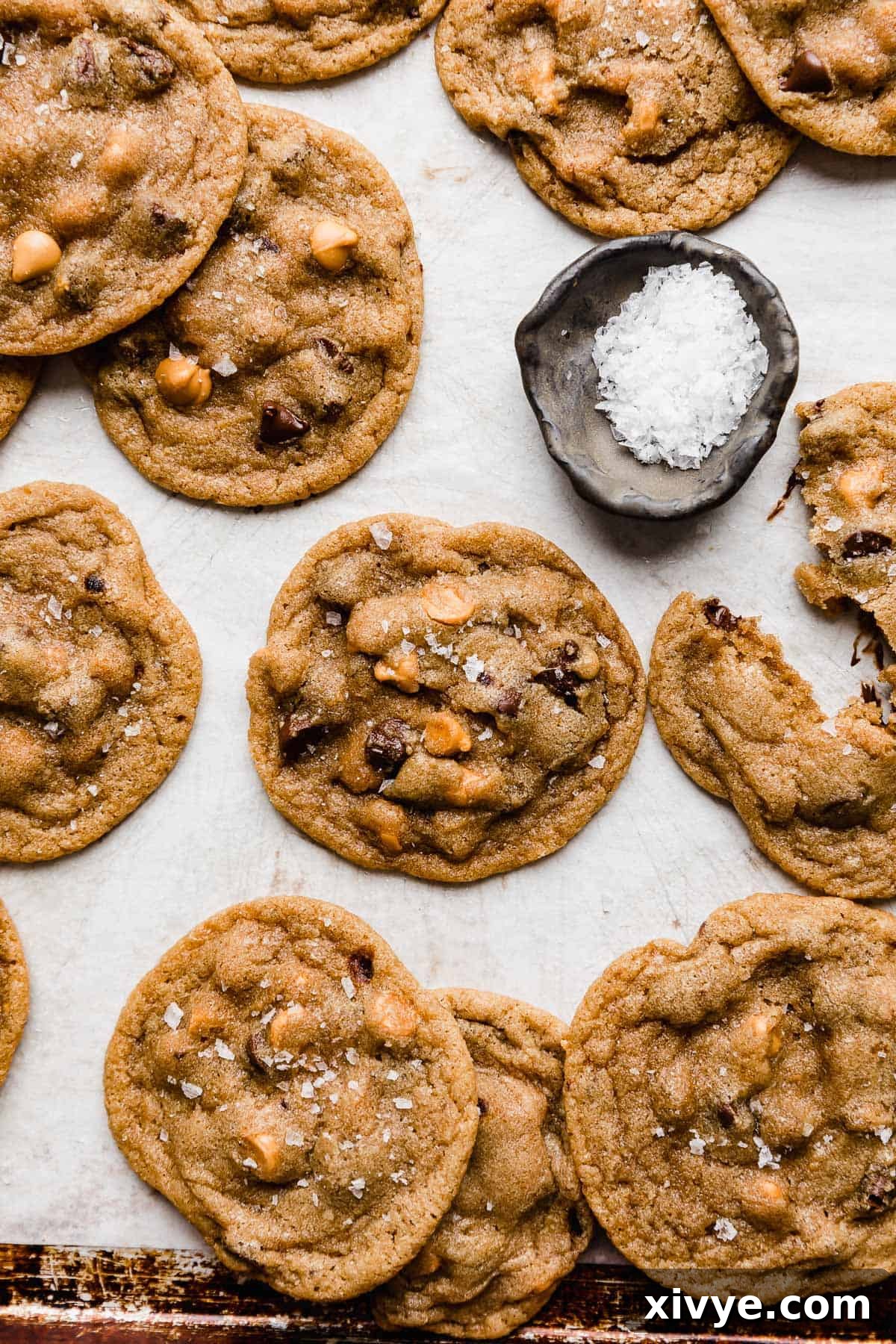 A batch of baked Butterscotch Chocolate Chip Cookies, topped with flaky sea salt, arranged on a white parchment-lined baking sheet.