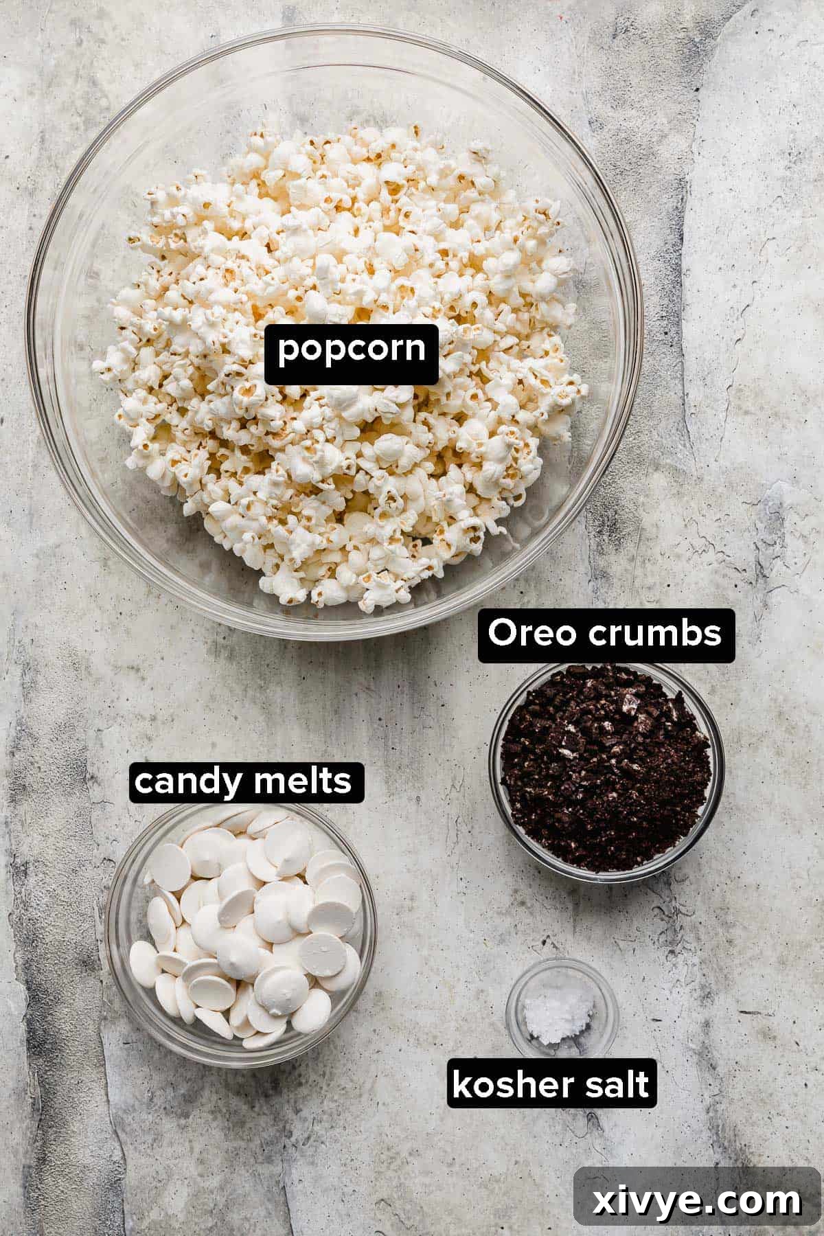 A flat lay showing the four simple ingredients for Oreo Popcorn: plain popped popcorn, white candy melts, crushed Oreo cookies, and a small dish of kosher salt.