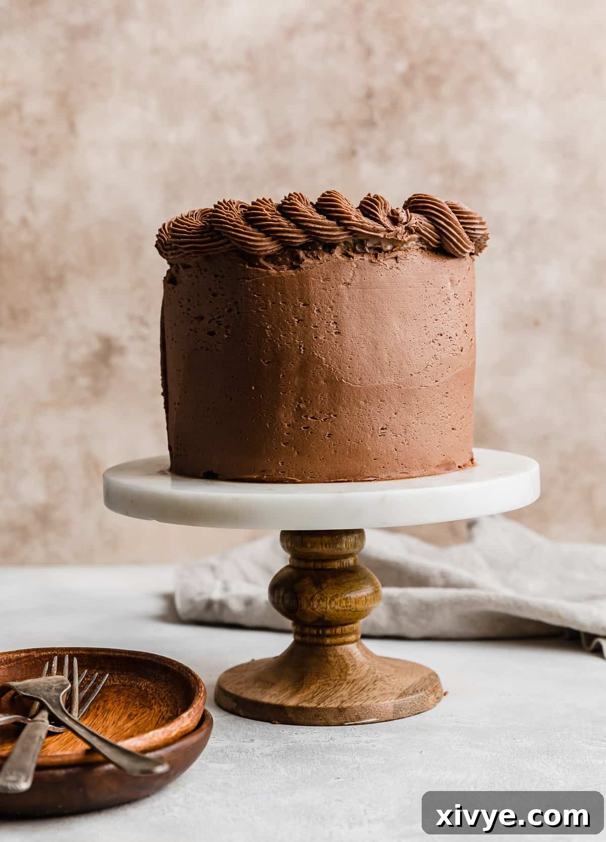 A beautifully frosted small three-layer German Chocolate Cake, adorned with smooth chocolate buttercream and a topping of coconut pecan frosting, presented on a cake stand against a warm, textured background.
