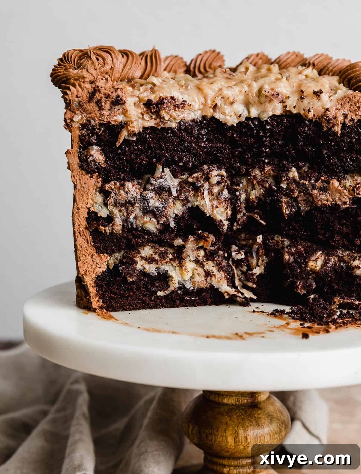 A magnificent three-layer Small German Chocolate Cake, expertly frosted and standing tall on a pristine white cake stand against a soft, light background, ready to be enjoyed.