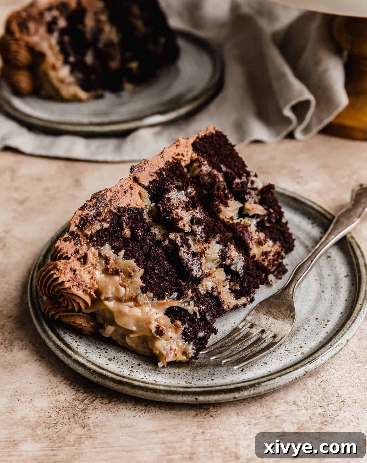 A generous slice of a stunning German Chocolate layer cake, showcasing its rich, dark chocolate cake and luscious coconut pecan filling, presented on a gray plate with a light brown background.